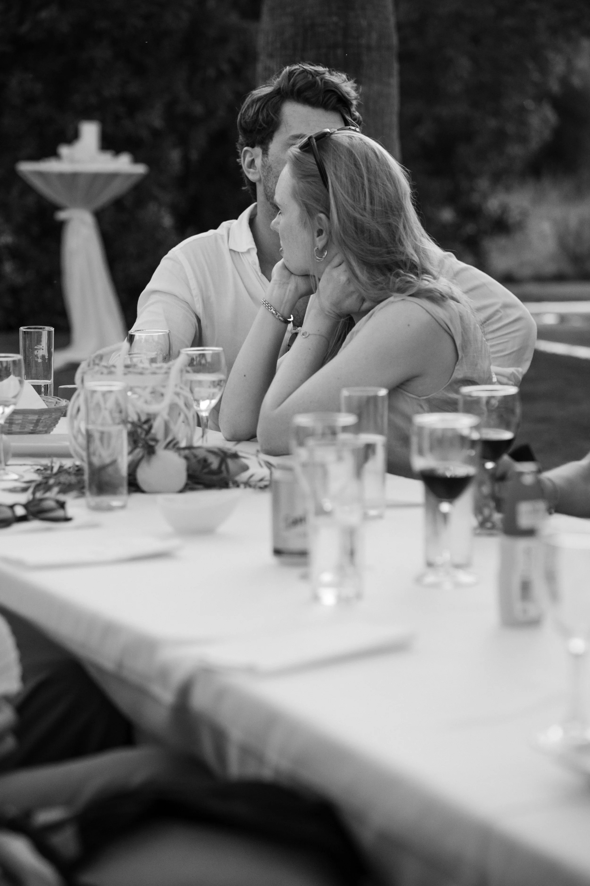 A black and white photo of a man and woman sitting closely at a table, engaged in an intimate moment, with various glasses and decorations on the table.
