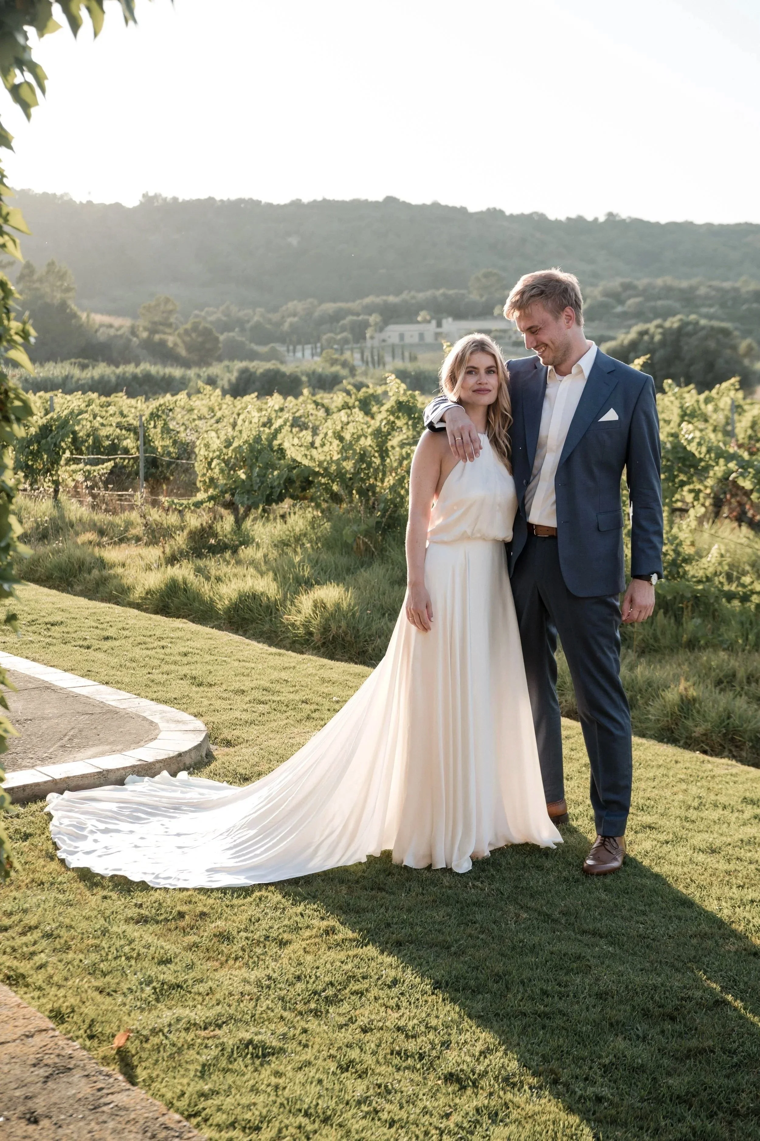 A bride and groom standing together outdoors in a vineyard. The bride is wearing a white strapless wedding gown with a long train, and the groom is in a navy blue suit with a white shirt. They are smiling and embracing each other, with the vineyard a