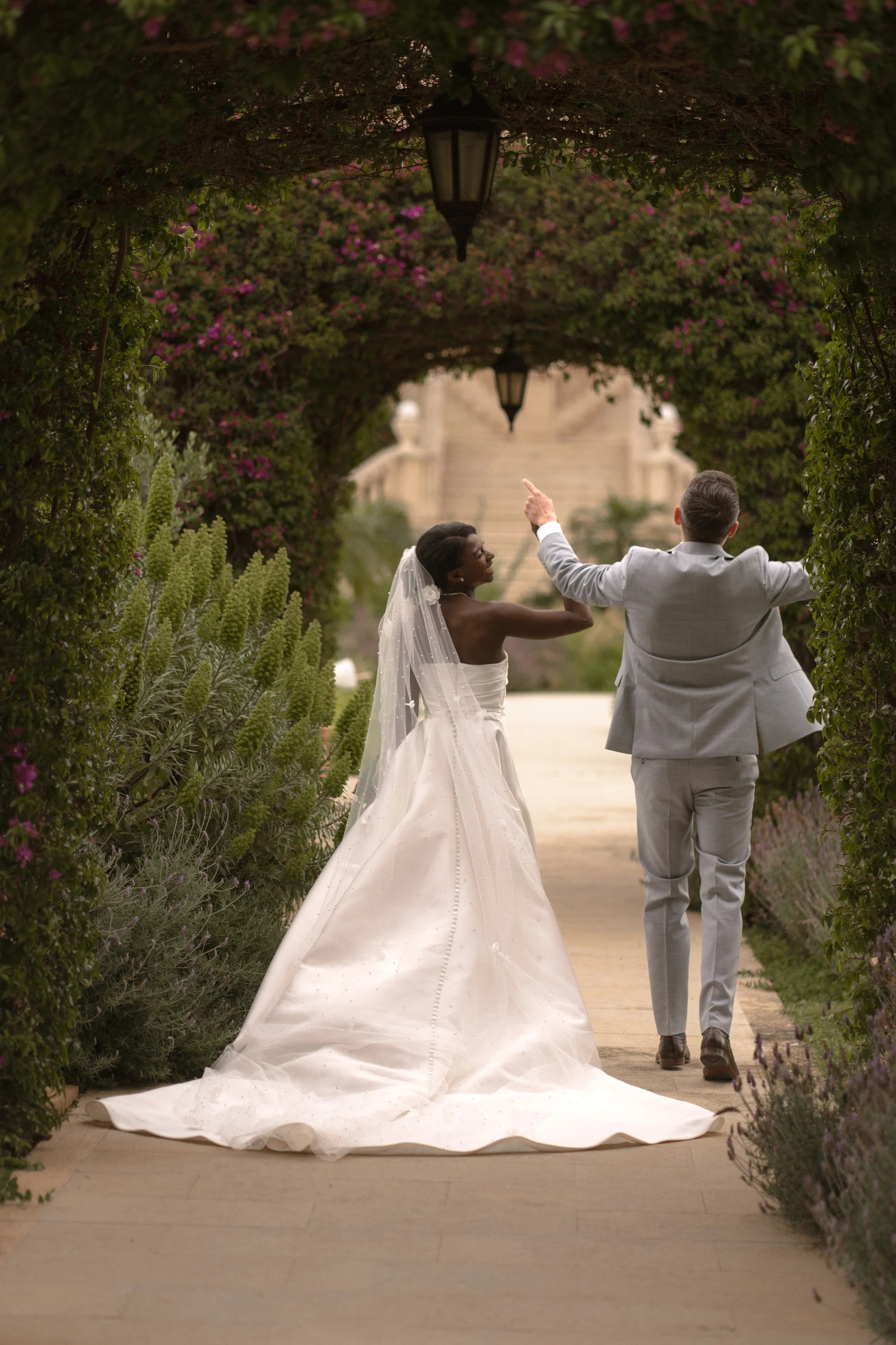 A bride and groom walk under a garden archway adorned with pink and purple flowers, with the groom pointing ahead and the bride smiling. The bride wears a white wedding gown and veil, and the groom wears a light gray suit. There are lanterns hanging 