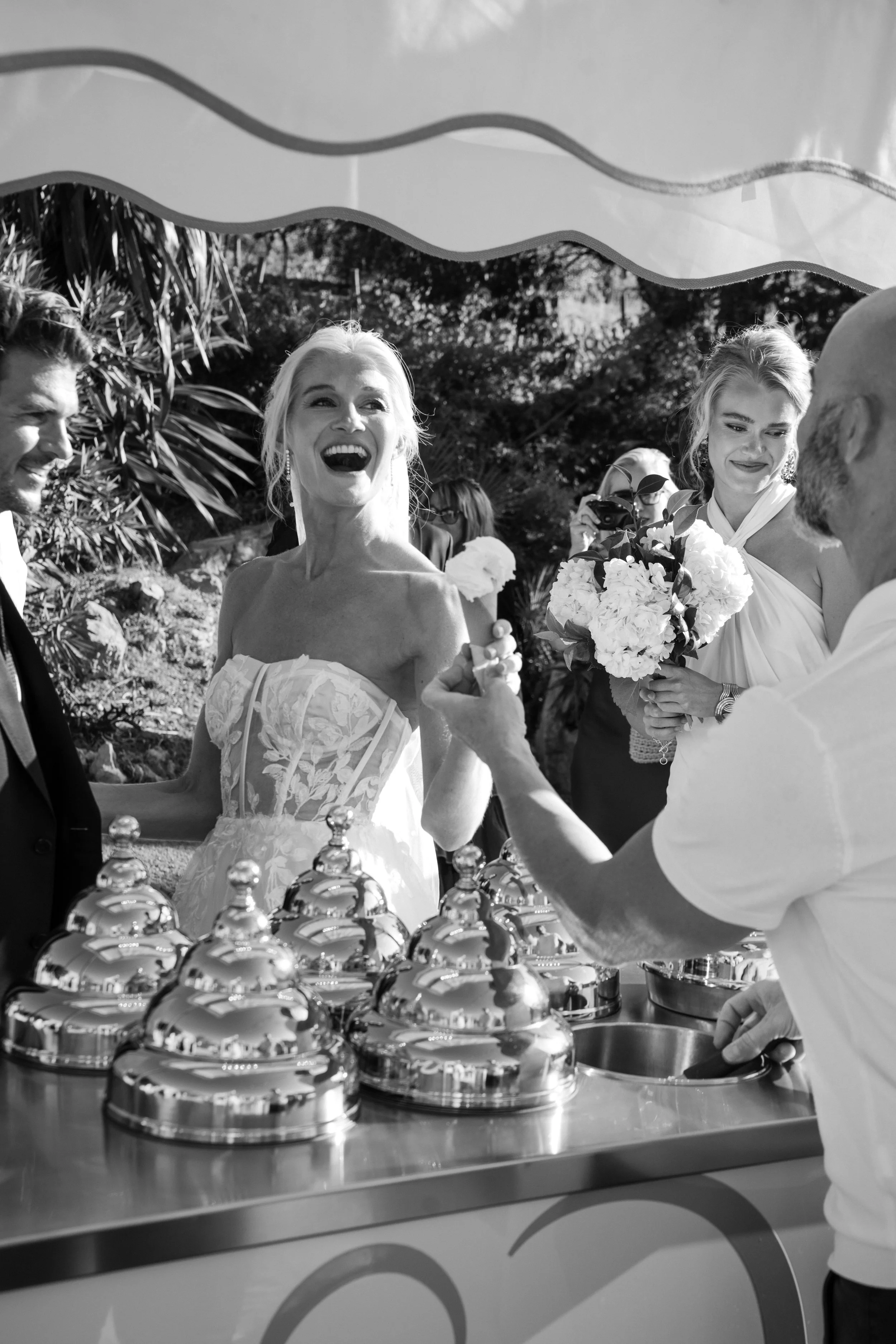 A black and white photo of a wedding celebration, with a bride and groom smiling at each other and surrounded by guests. The bride holds a bouquet, and there is a table with shiny domed trays, possibly for serving food or desserts.