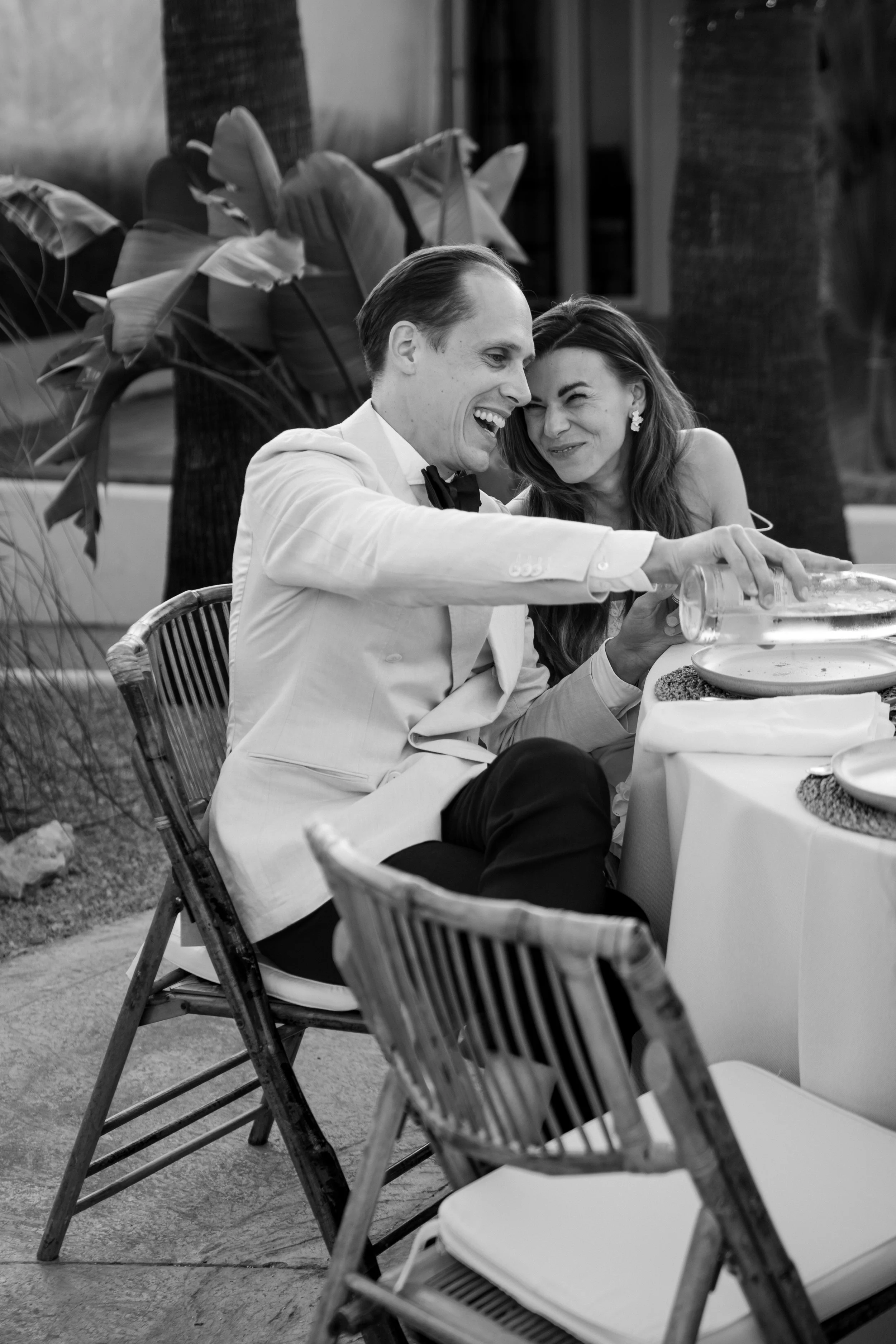 A black and white photo of a man and woman sitting at a table outdoors, smiling and laughing together. The man is wearing a tuxedo and the woman has long hair and earrings. There are chairs and plants in the background.