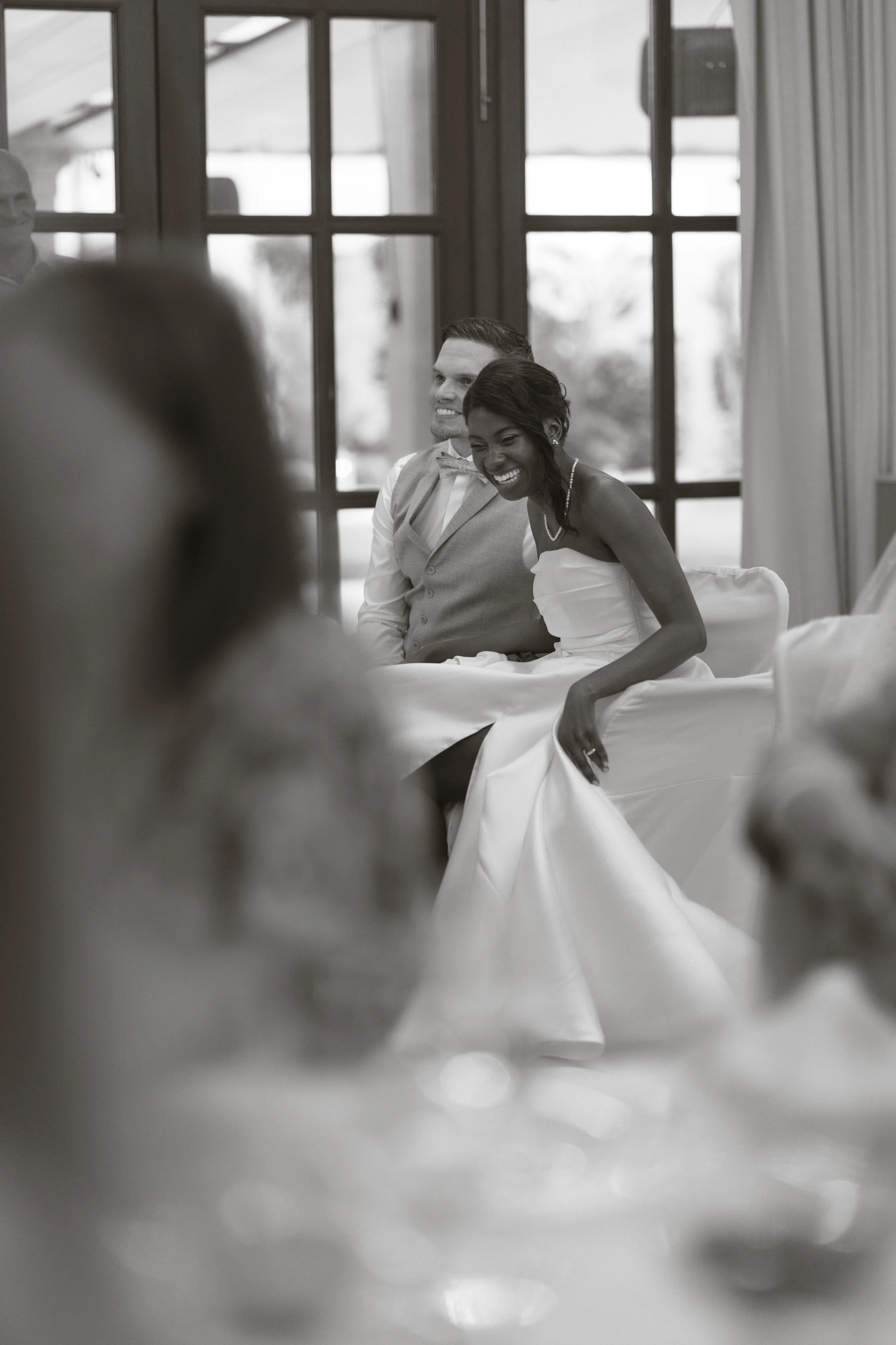 A black and white photo of a bride and groom seated inside a room with large windows, smiling and looking at each other, during a wedding celebration.