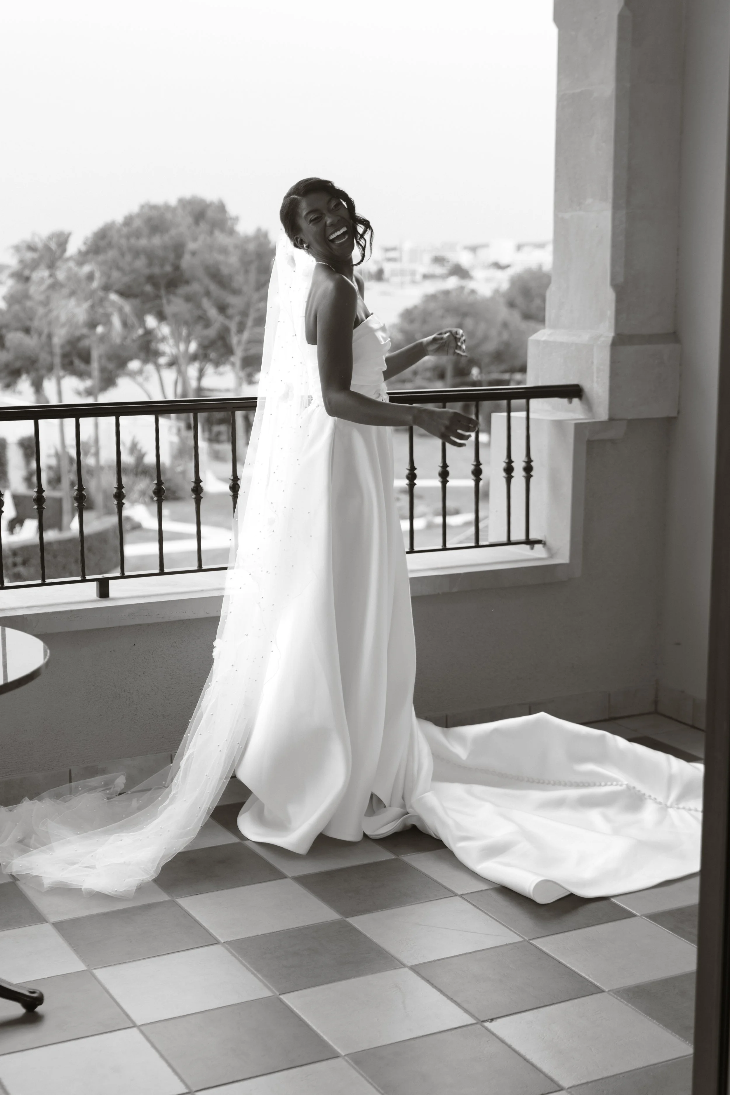 A joyful bride in a strapless wedding gown and veil, standing on a balcony with a view of trees and buildings in the background, smiling and laughing.