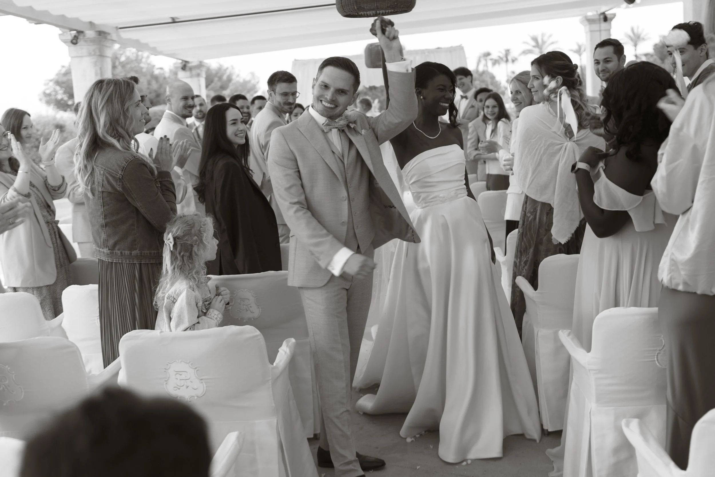 A young man in a suit and bow tie holding a large bell above his head at a wedding reception, surrounded by smiling guests clapping and celebrating outdoors under a pavilion with palm trees in the background.