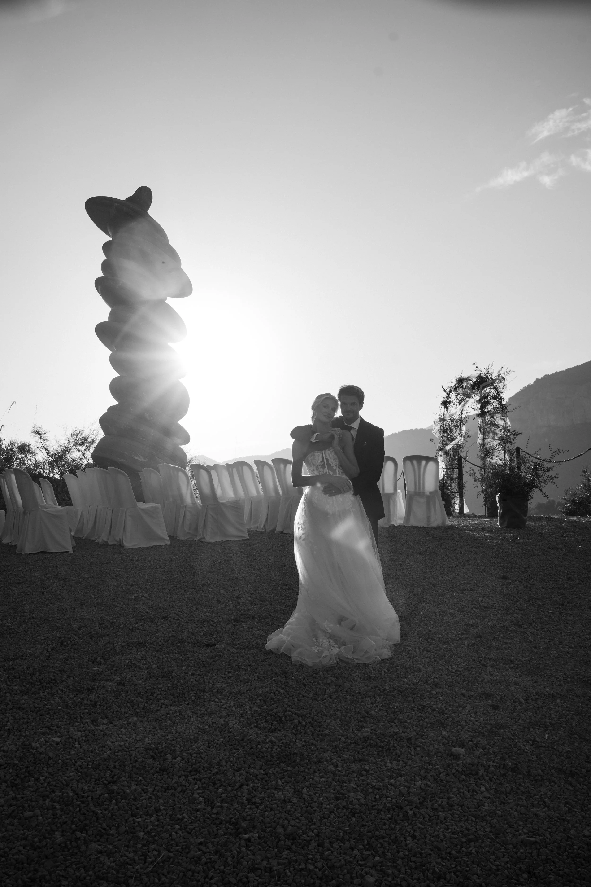 A couple, dressed in wedding attire, stands outdoors during sunset. The bride wears a flowing gown, and the groom is in a suit. Behind them are empty chairs draped with fabric, and decorative balloon sculptures are visible nearby. The scene is set ag