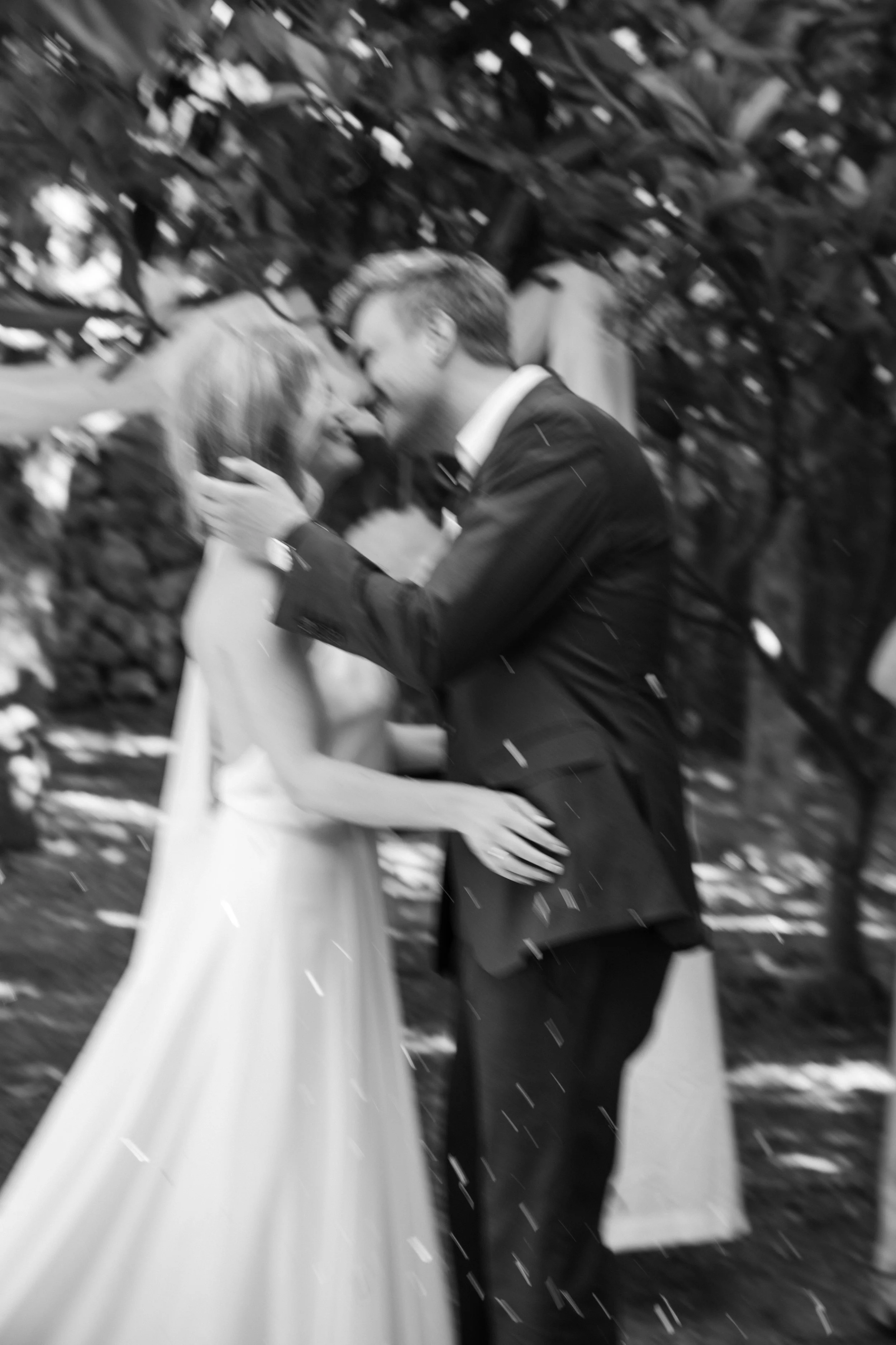 A couple is sharing a kiss during a wedding ceremony outdoors, with a blurred background of trees and falling rain.