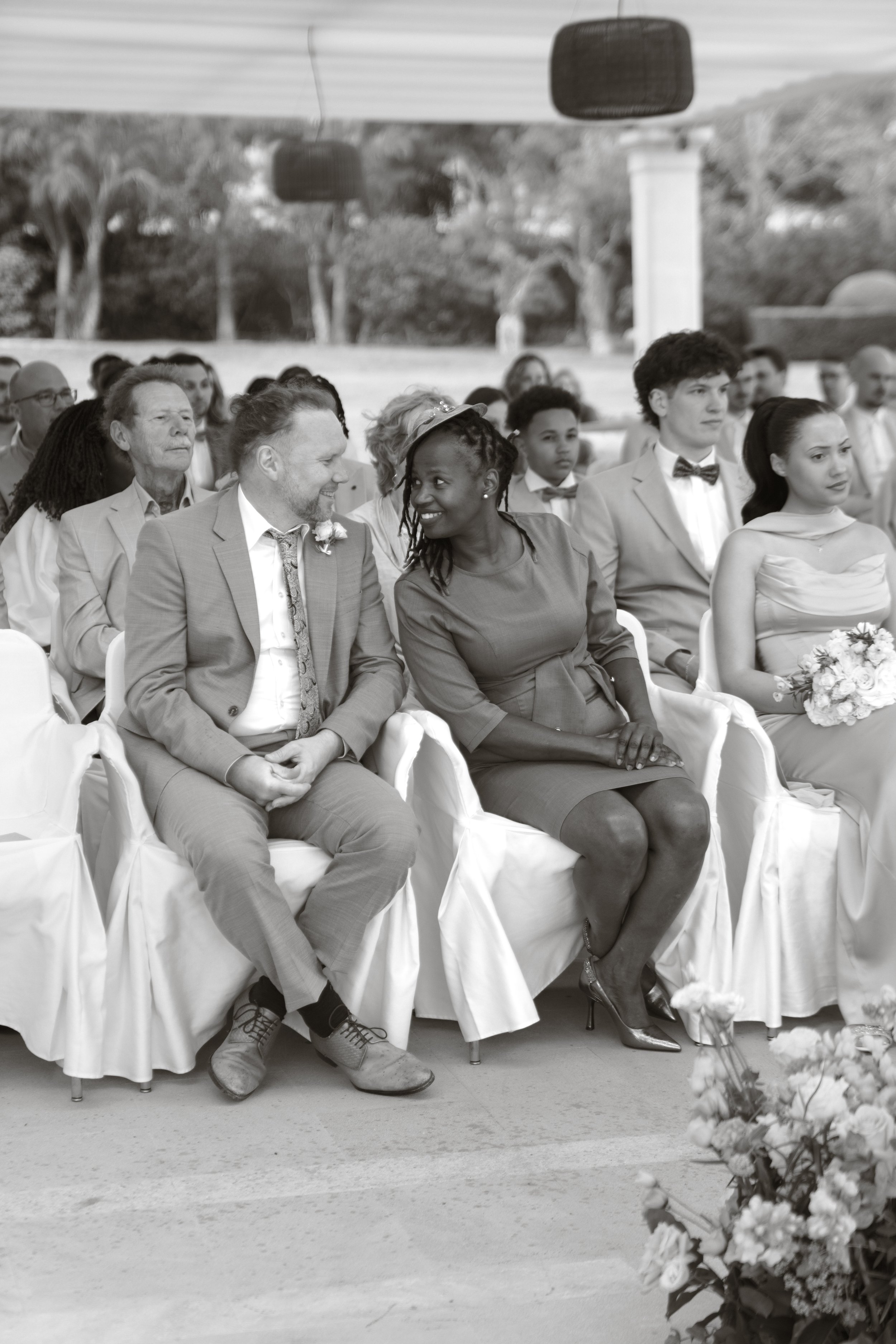 A diverse group of people sitting at an outdoor wedding ceremony, dressed in formal attire, with a flower arrangement in the foreground.