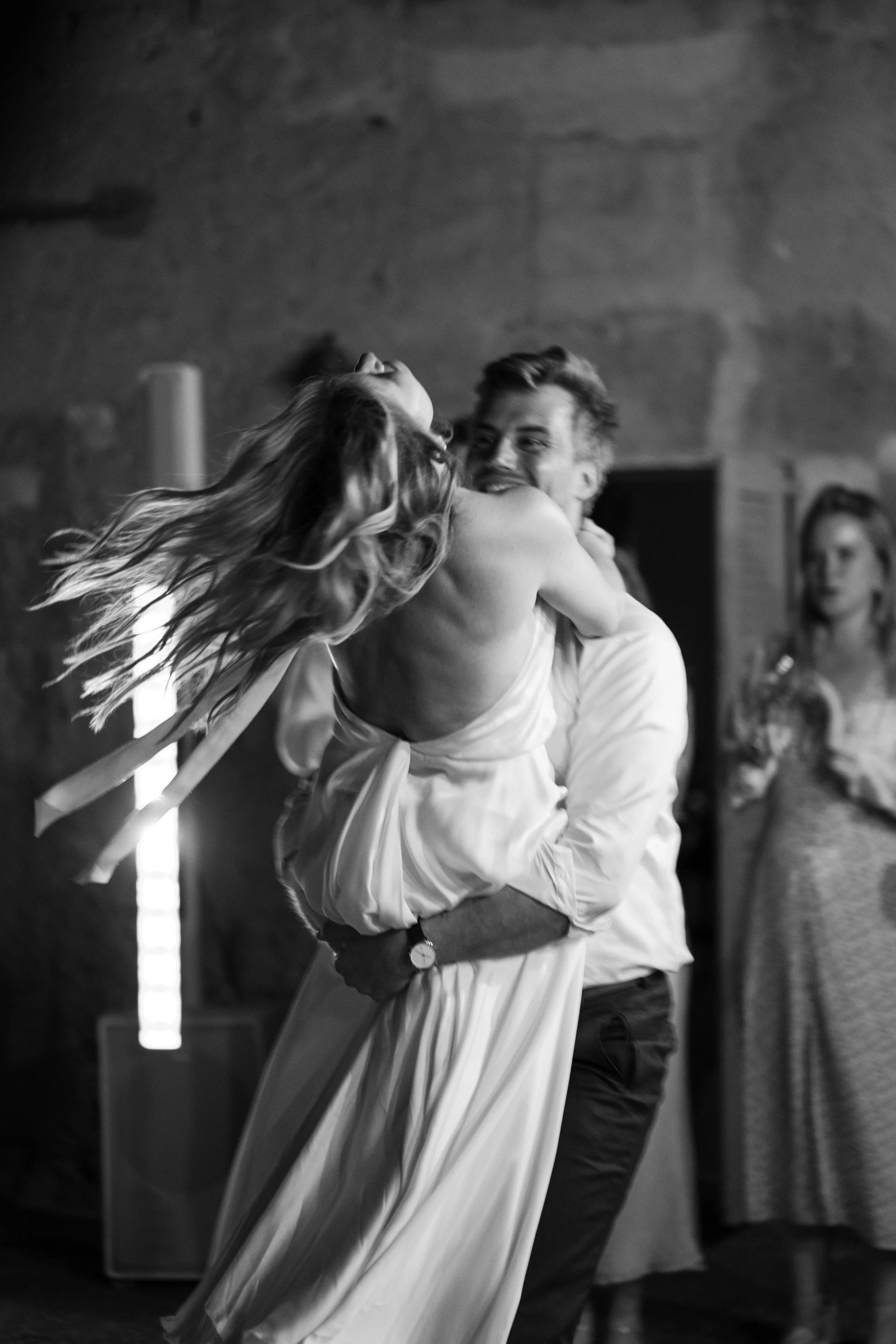A couple dancing closely, with the man smiling and the woman leaning back with her hair flowing, in a dimly lit space.
