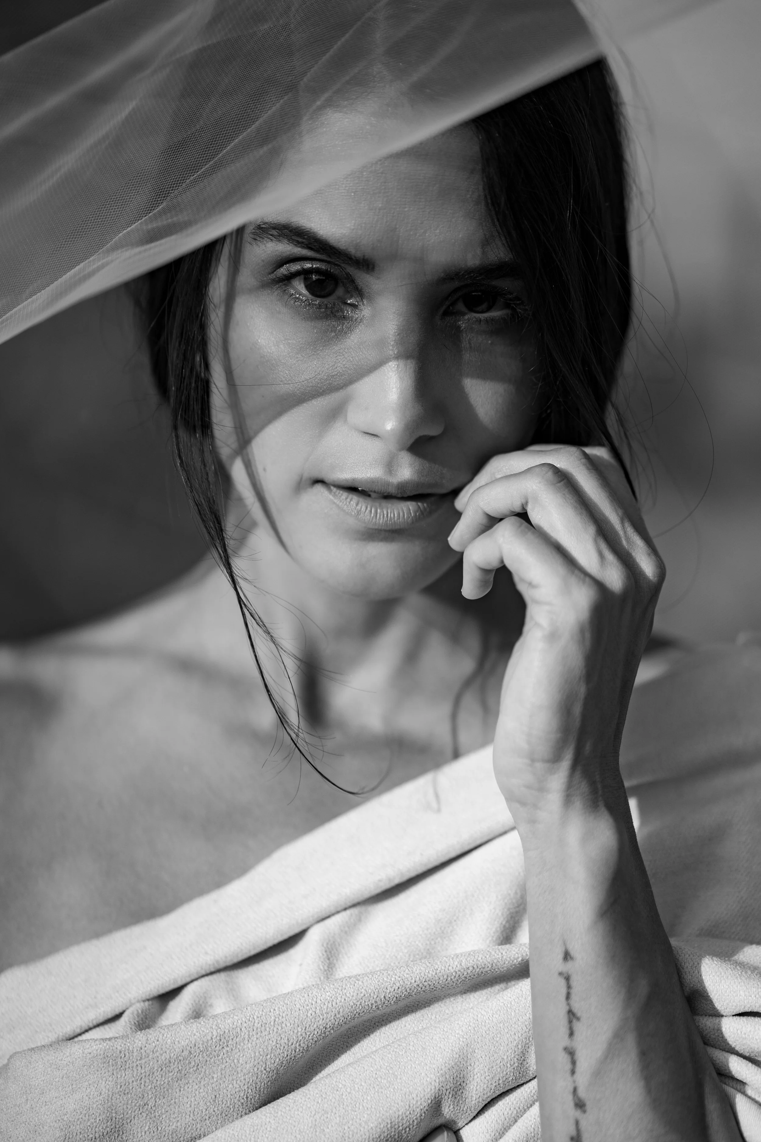 A woman with dark hair and visible tattoo on her arm, wearing a wide-brimmed hat and a light-colored off-shoulder top, gazing at the camera with her hand near her face.