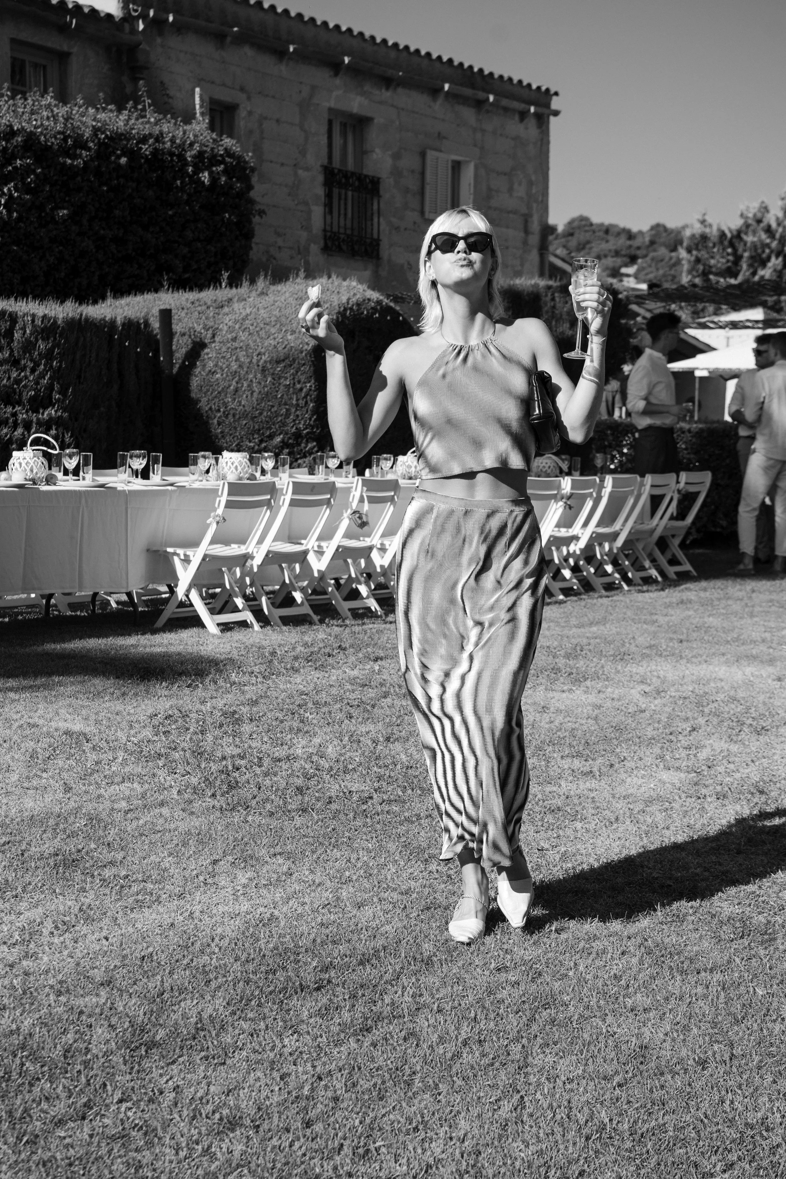 A woman in stylish clothing and sunglasses is walking on a grassy lawn, holding a drink in a glass and a small snack in her hand. In the background, there is a long table set up for an outdoor event with chairs and decorative items, and a group of pe