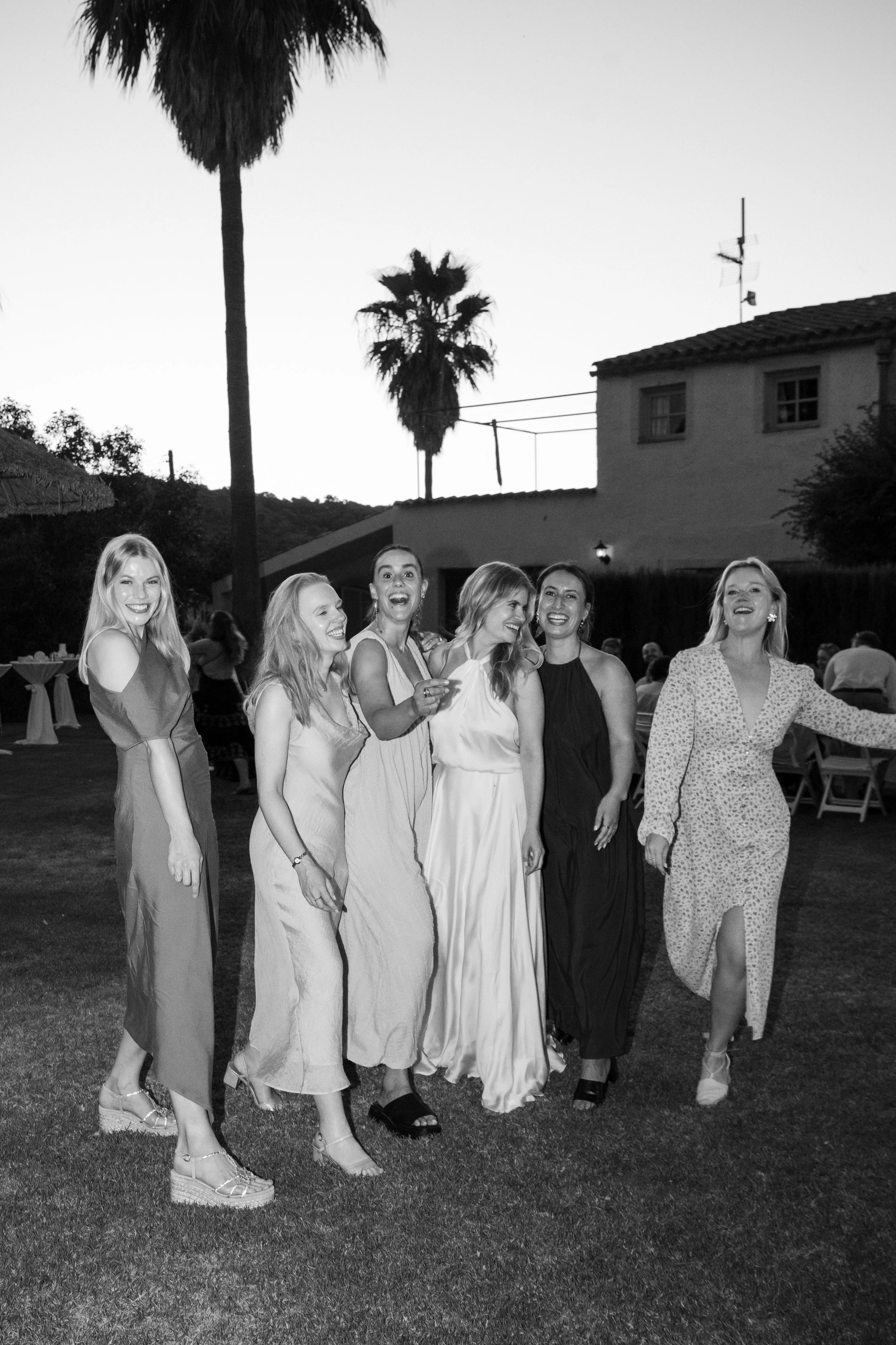A group of seven women smiling and walking outdoors during evening with palm trees in the background, dressed in elegant attire.
