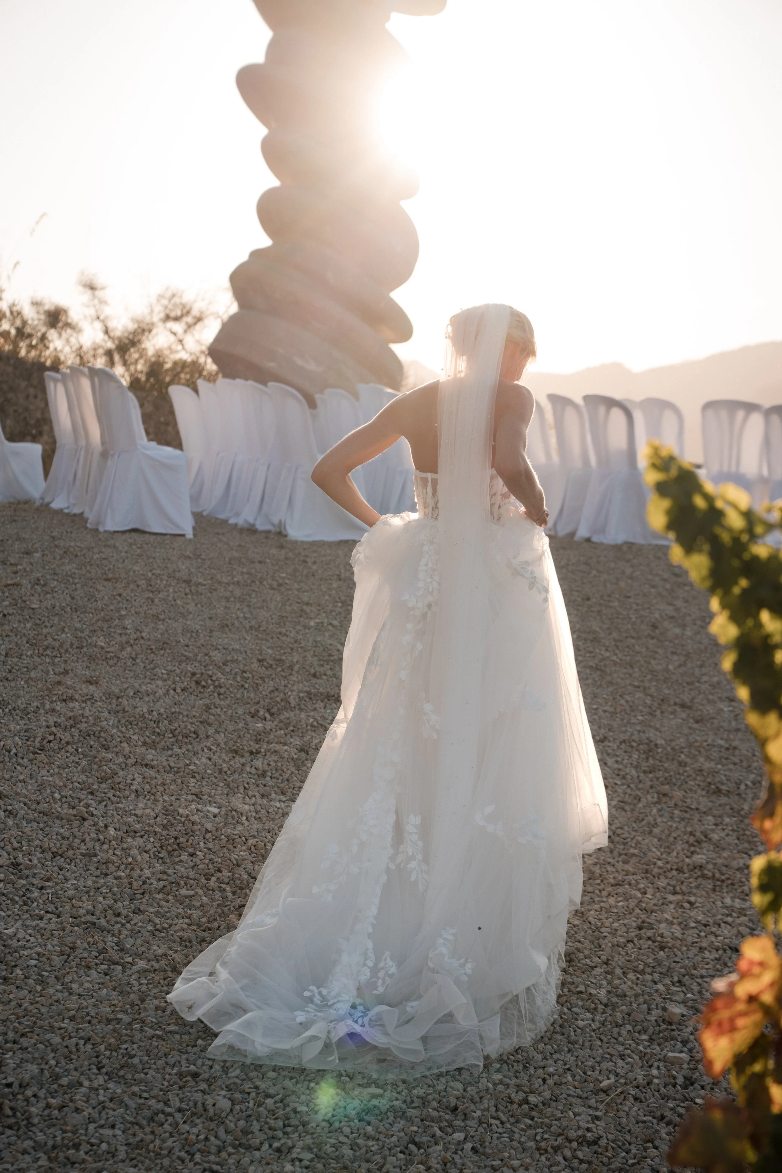 A woman in a white wedding dress standing outdoors on gravel with her back to the camera, facing a large stacked stone sculpture, with chairs arranged in a semi-circle behind her, and the setting sun creating a bright glow.
