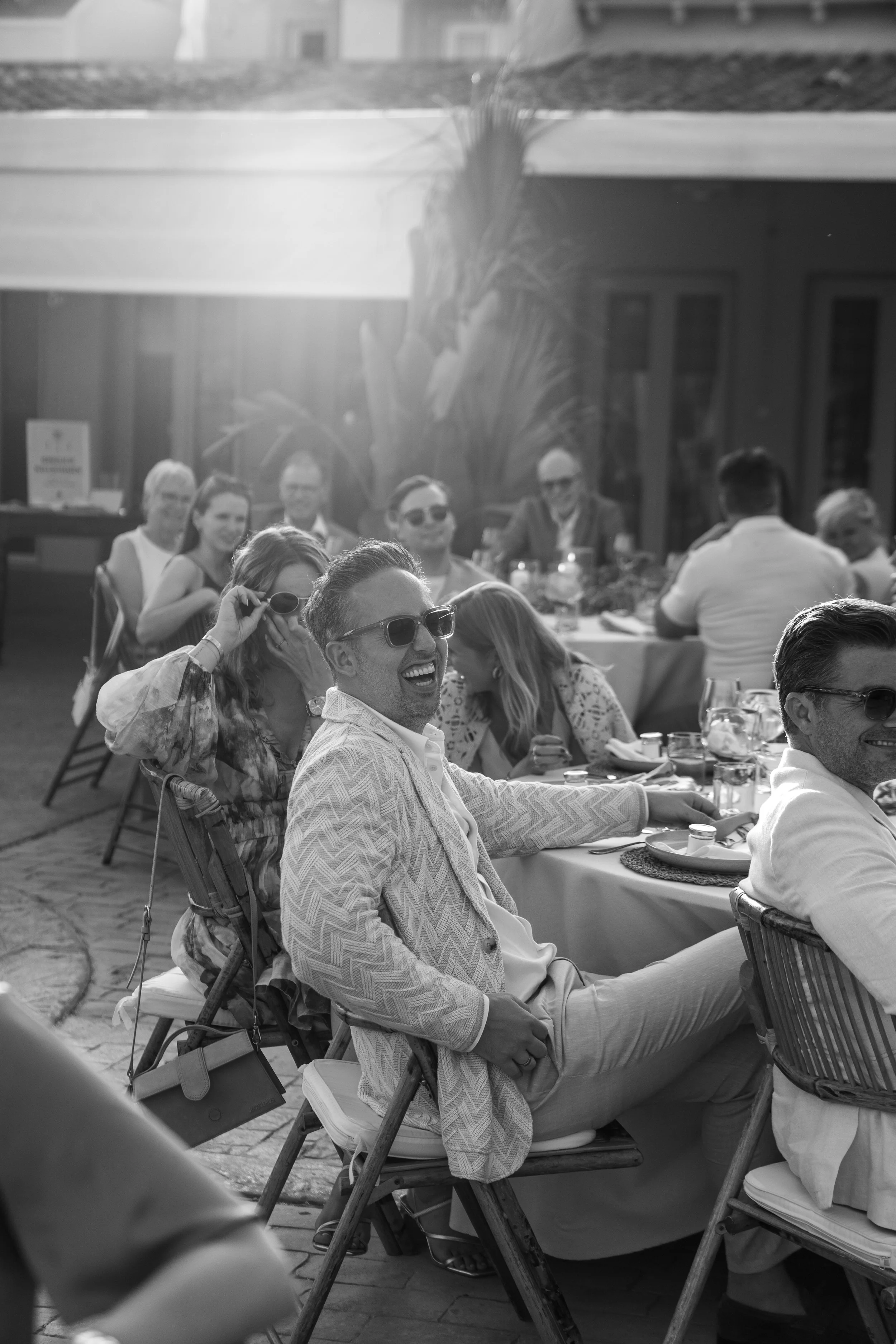 People having a meal outdoors at a sunny gathering, some wearing sunglasses, smiling and engaging in conversation.
