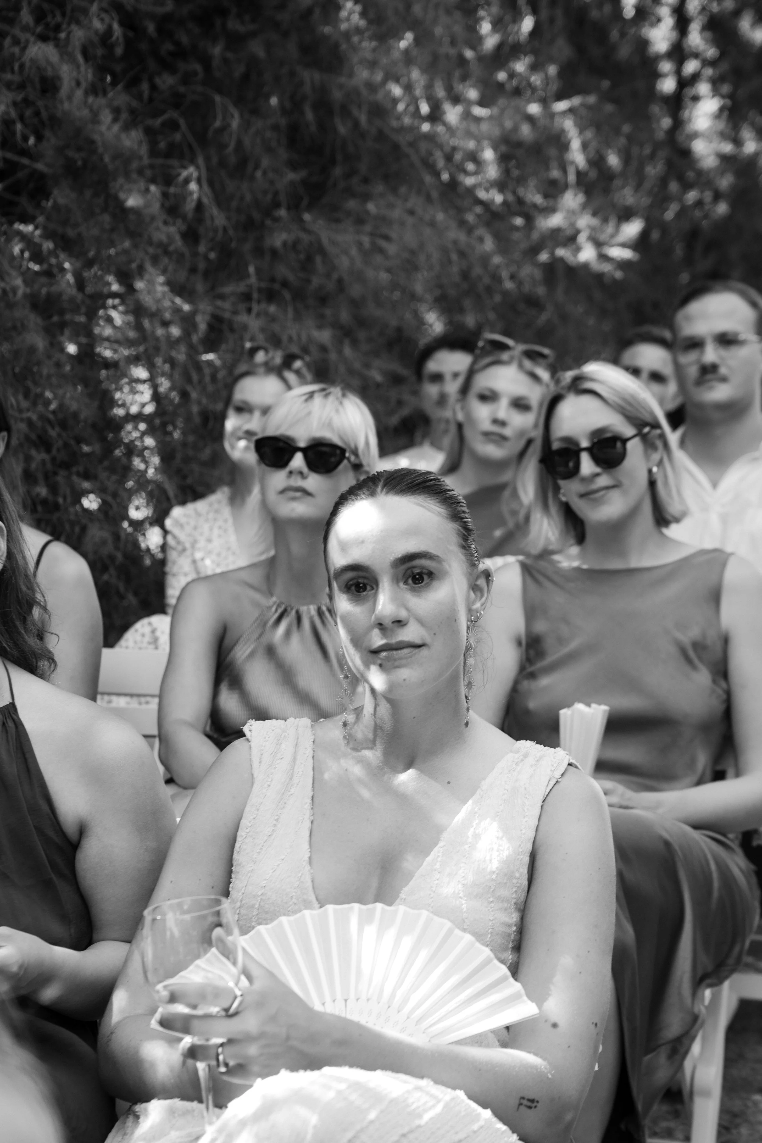 A group of women and men at an outdoor gathering, some wearing sunglasses, with trees in the background. The focus is on a woman with earrings and a sleeveless top, holding a paper fan and a glass, with others standing around her.