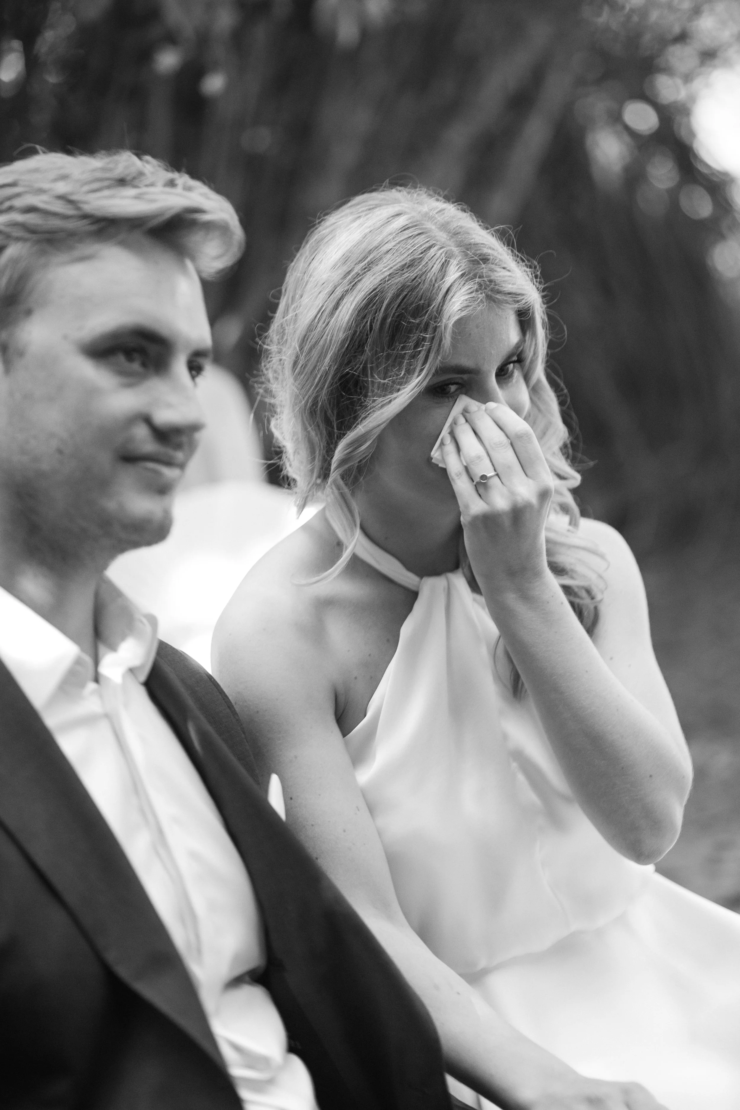 A black-and-white photo of a man and woman seated side by side. The man is dressed in a suit and bow tie, smiling slightly, while the woman appears to be wiping tears from her eye, wearing a sleeveless dress. The background is blurred with trees, sug