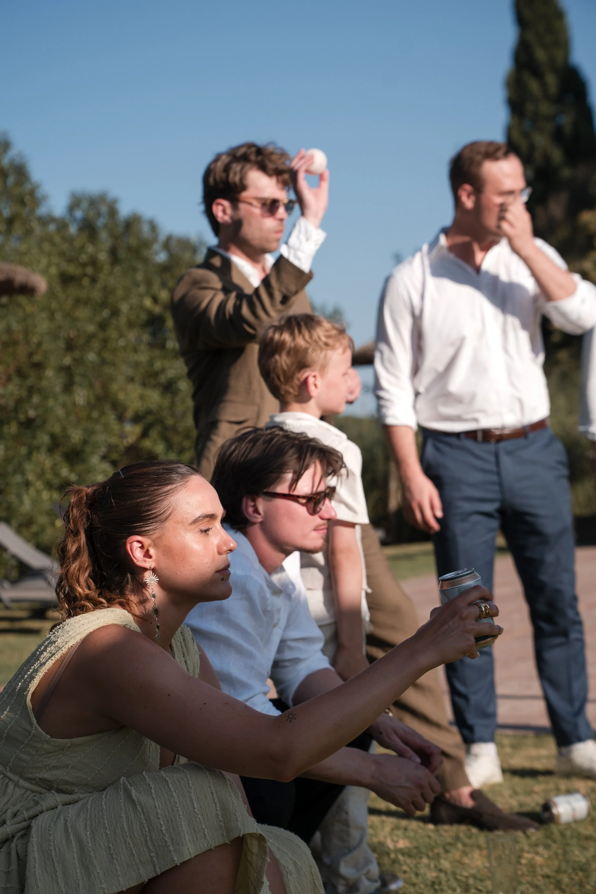 Group of people sitting outdoors, with one woman in the foreground meditating and others standing or sitting behind her, against a backdrop of trees and a clear sky.