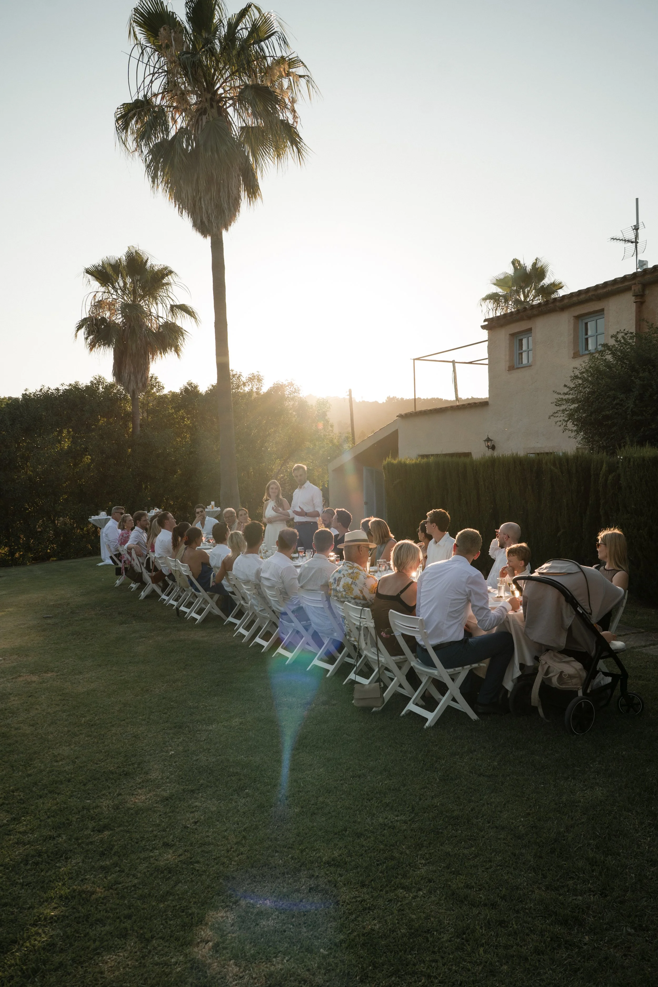 A large outdoor gathering at sunset with guests seated at long white tables, some standing and talking. Two people are standing and possibly giving a toast. The setting features palm trees, a house, and green shrubbery in the background.
