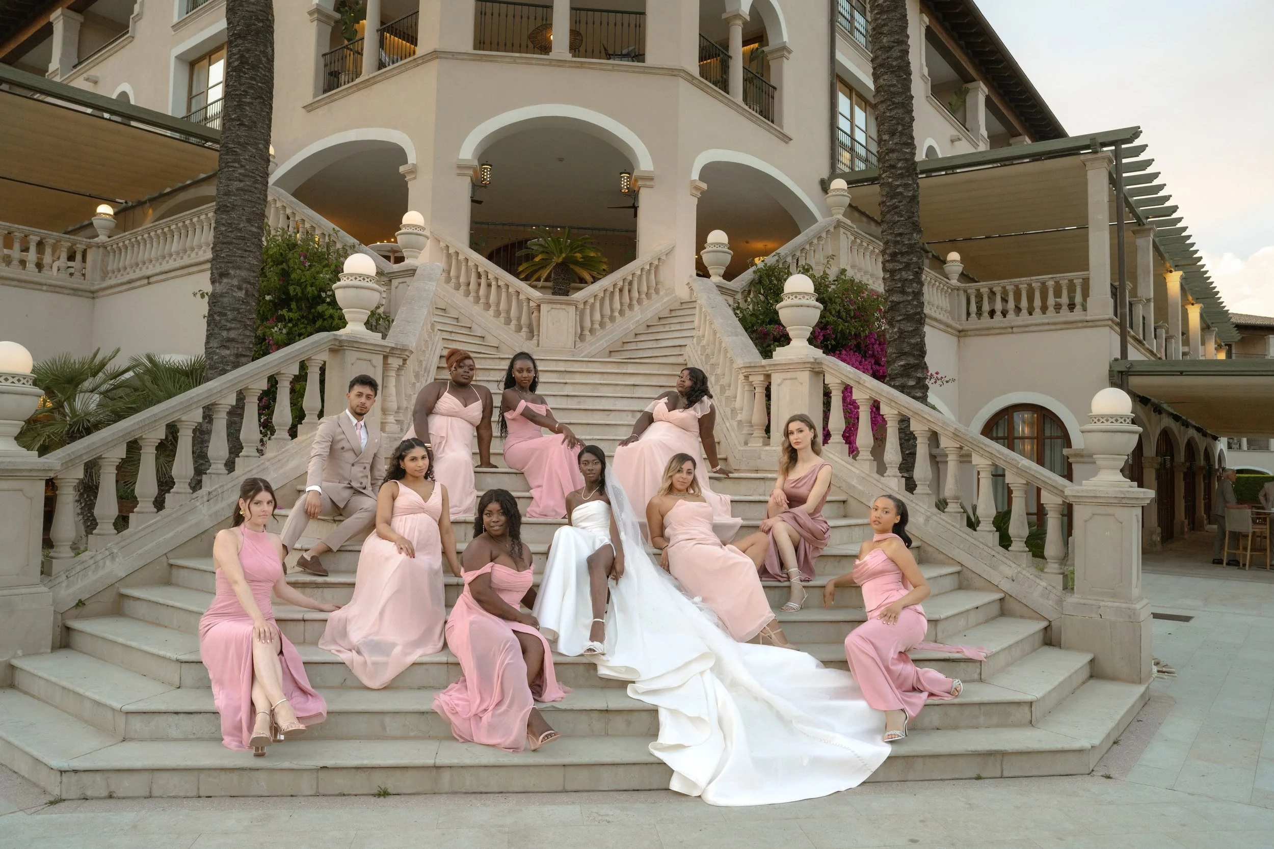 A group of people, mostly women in pink dresses and one man in a beige suit, sitting and standing on a grand staircase outside a large, elegant building with arches and balconies, during daylight.
