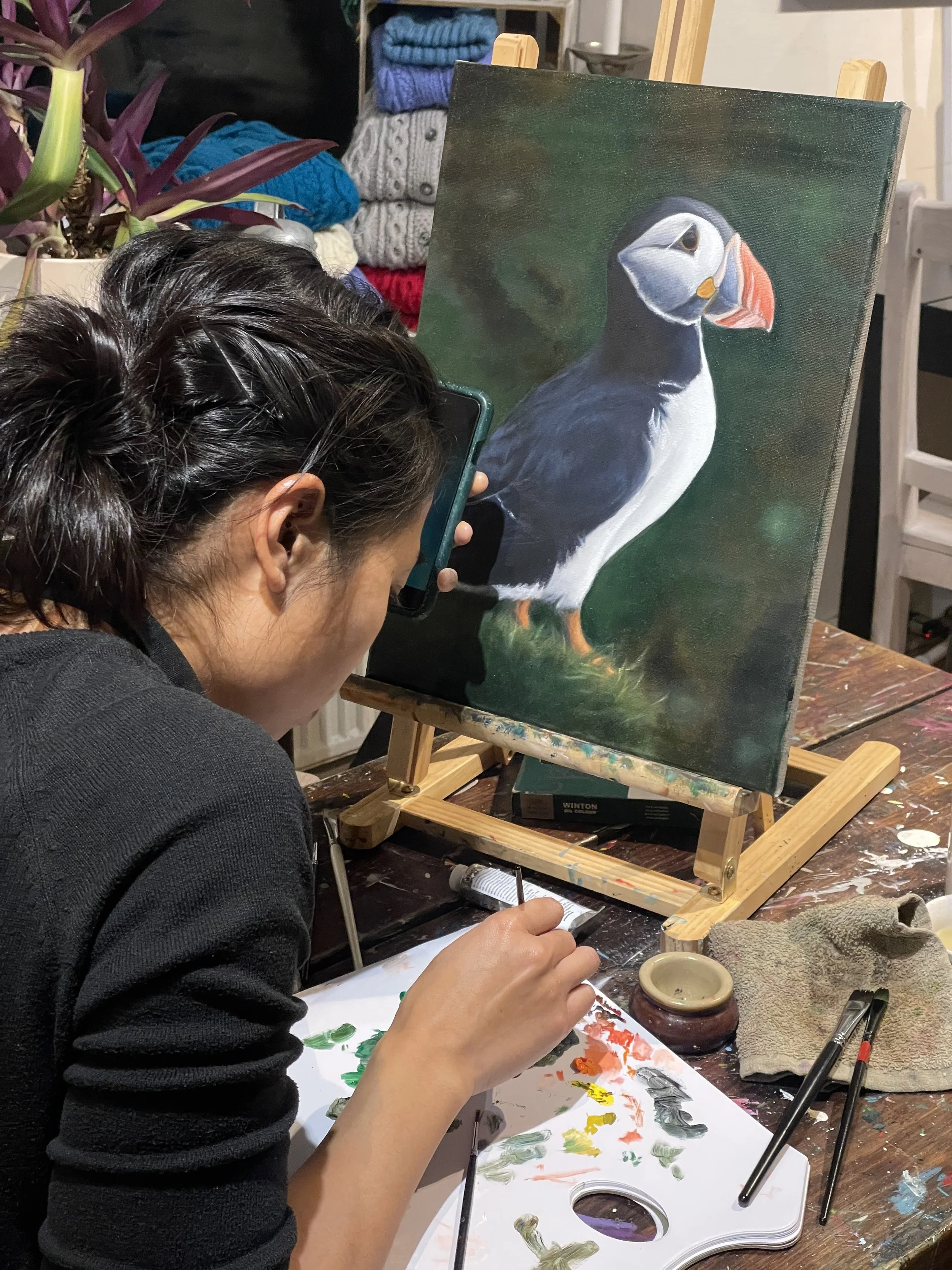 A woman is painting a detailed portrait of a puffin bird on canvas, sitting at a cluttered art table with various brushes, paints, and a palette. In the background, there are neatly folded sweaters and a potted plant with purple and green leaves.