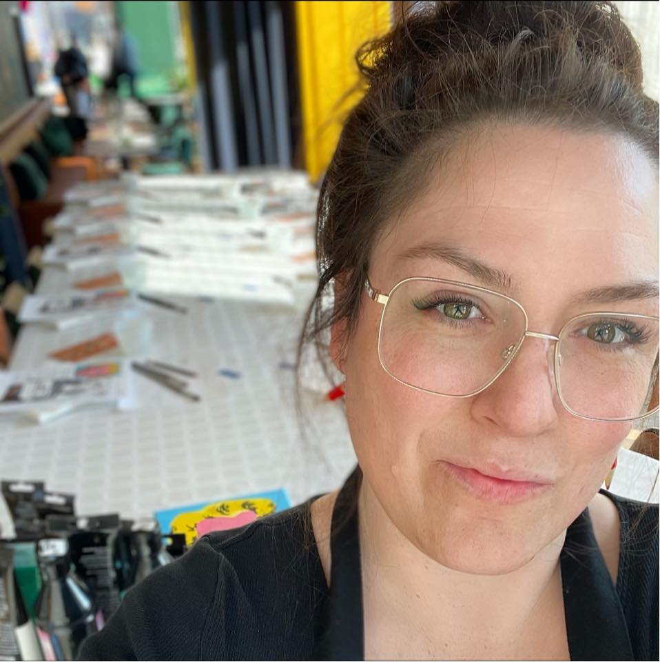 A woman with glasses and brown hair tied back, smiling at a table filled with books and magazines in a room with yellow and green walls.
