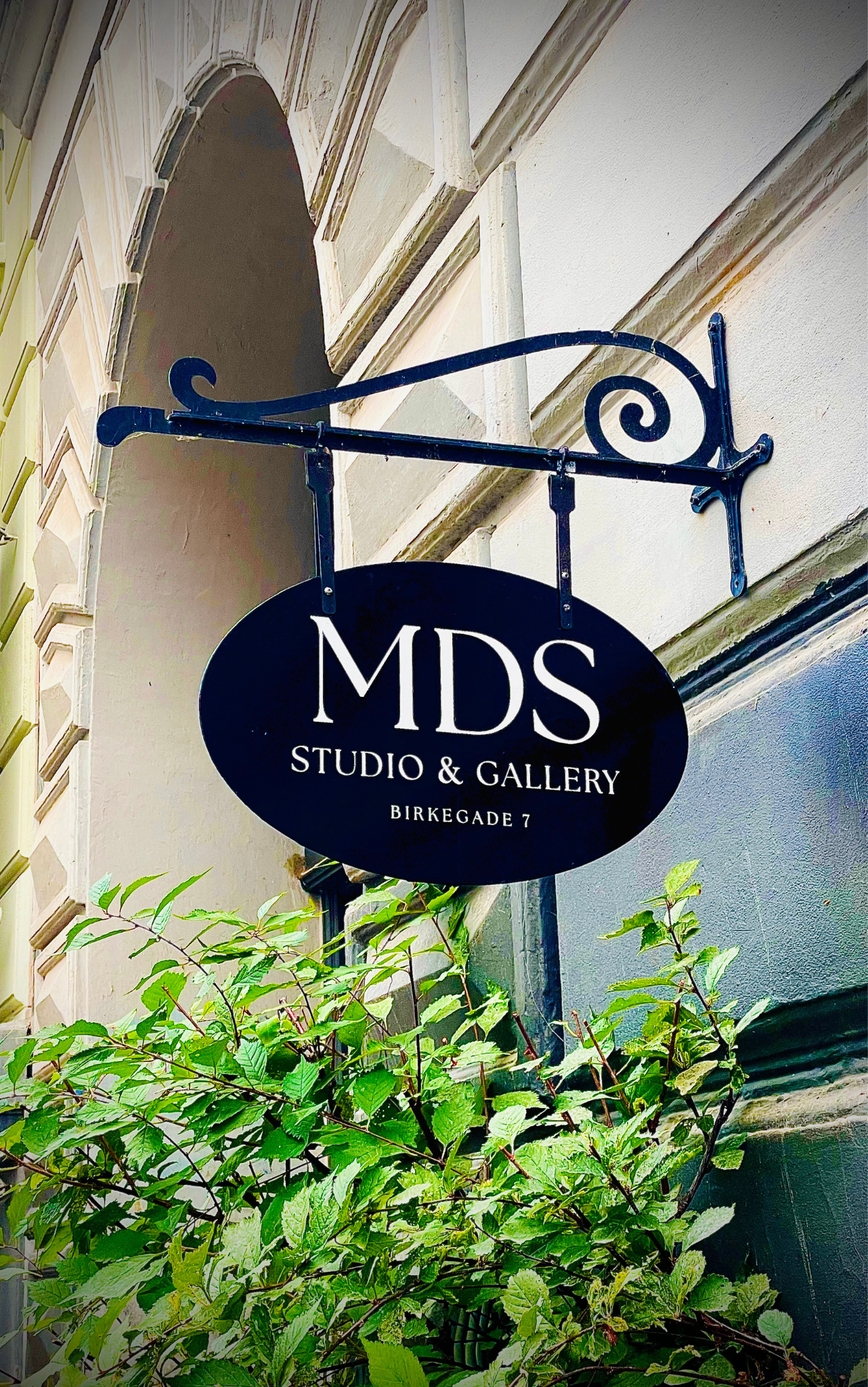 Black oval sign hanging from a decorative metal bracket reads 'MDS Studio & Gallery Birkegade 7'; the sign is outdoors next to a building with beige stone and white walls, with green leafy plants in front.