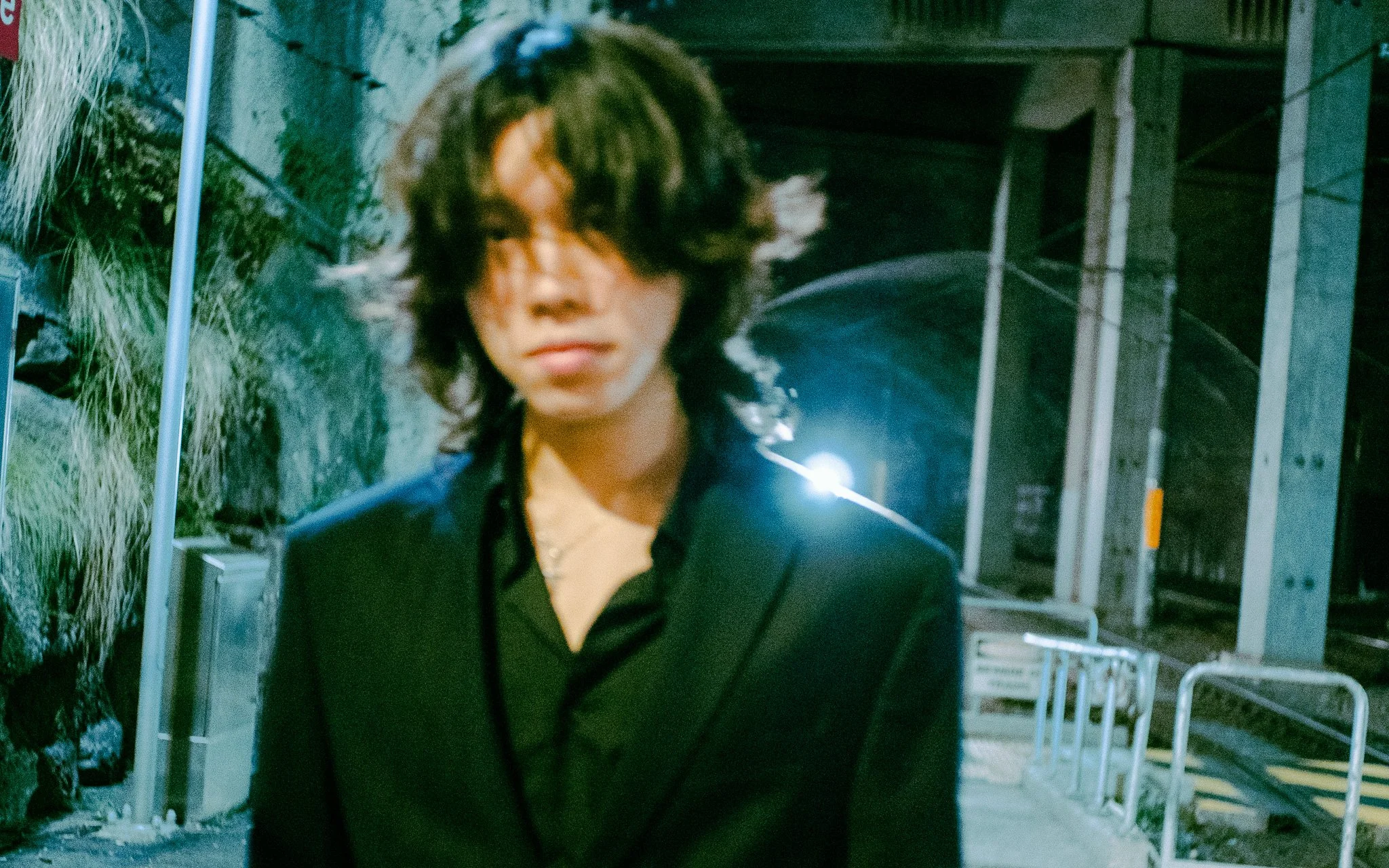 A man with dark, curly hair in a black shirt walking outside at night, with a background of building structures, a chain-link fence, and greenery.