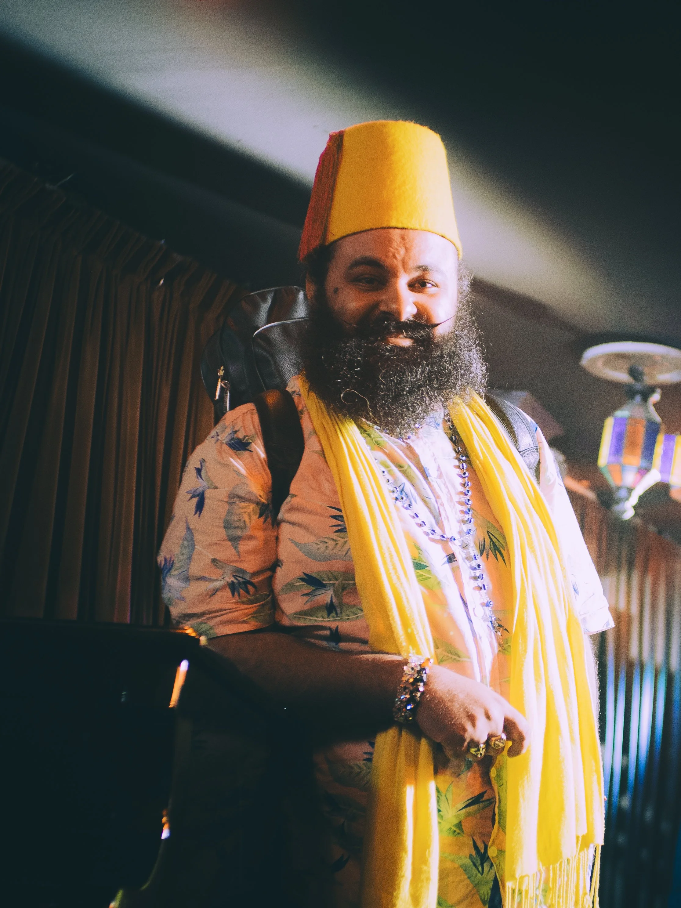 A man with a large beard and mustache wearing a yellow hat and shirt with a tropical leaf pattern, holding a yellow scarf or sash, and smiling at the camera inside a dimly lit room with a dark curtain in the background.