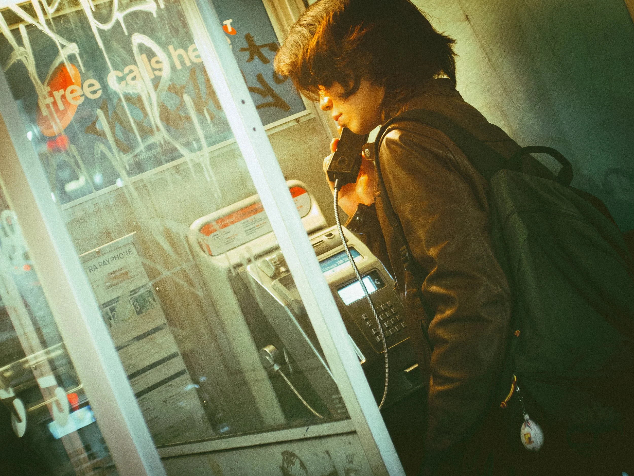 A person with brown hair, wearing a black leather jacket, is at an ATM machine, holding a phone to their ear. The ATM has visible buttons and a screen, with graffiti on the wall beside it.