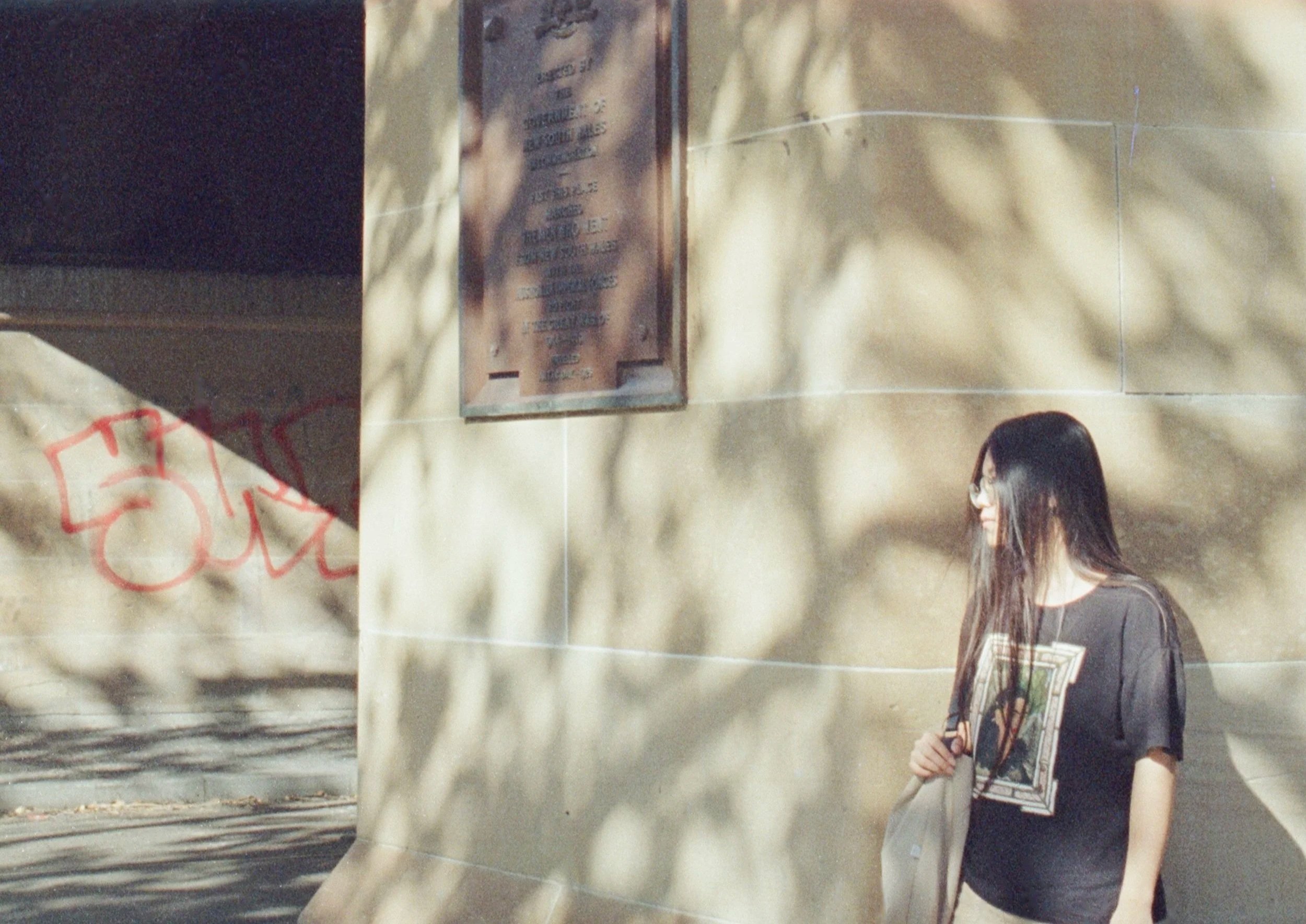 A woman with long dark hair and glasses, walking outdoors next to a reflective building wall with shadows of tree leaves. A metal sign and graffiti are visible on the wall.
