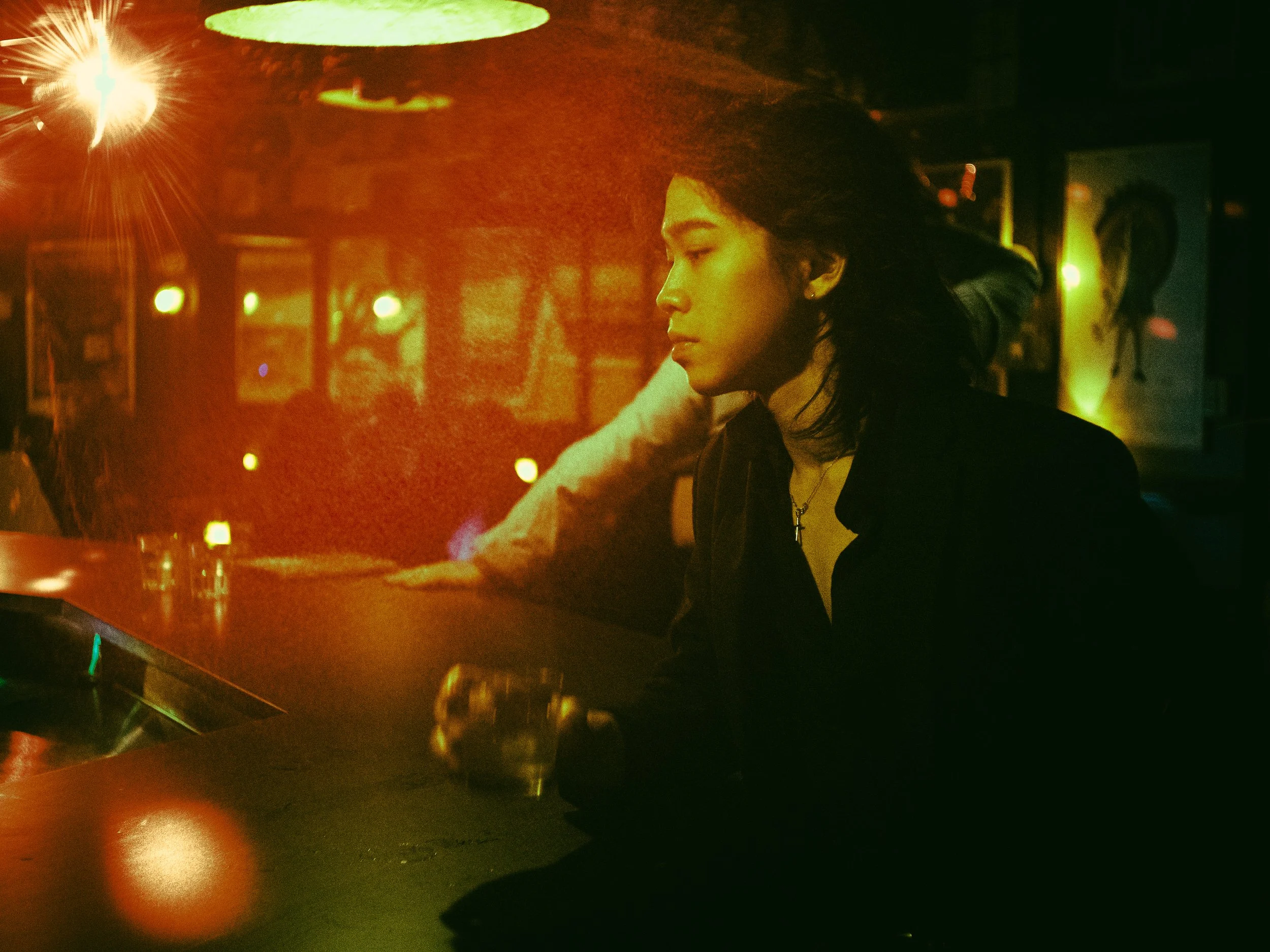 Young man with dark hair and earrings resting her chin on her hand, sitting at a bar with colorful glowing lights in the background.