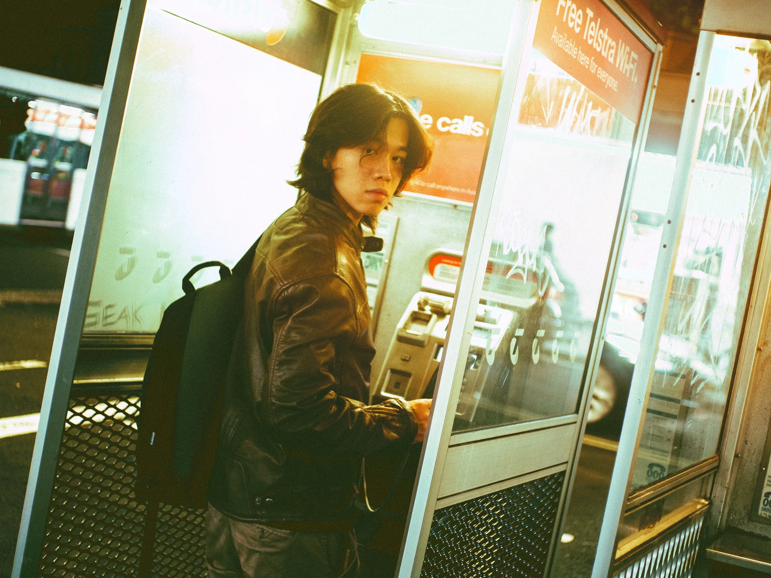 A young man with shoulder-length hair, wearing a brown leather jacket and carrying a backpack, sitting in front of a glass-walled telephone booth at a convenience store or gas station.