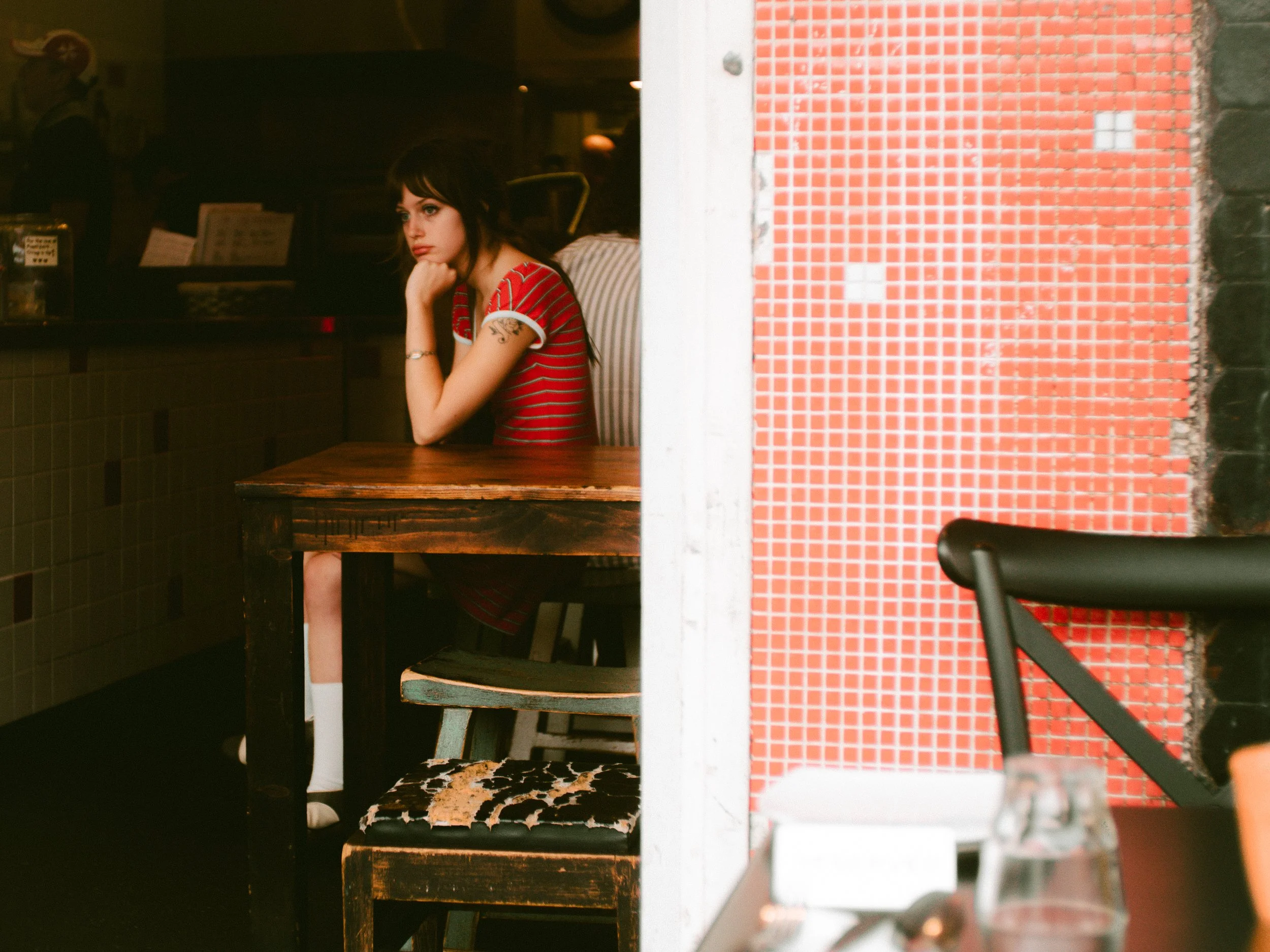 A young woman wearing a red striped shirt sitting alone at a restaurant table, resting her chin on her hand, looking pensively to the side. The restaurant is dimly lit, with a mixture of red tile and brick on the walls.