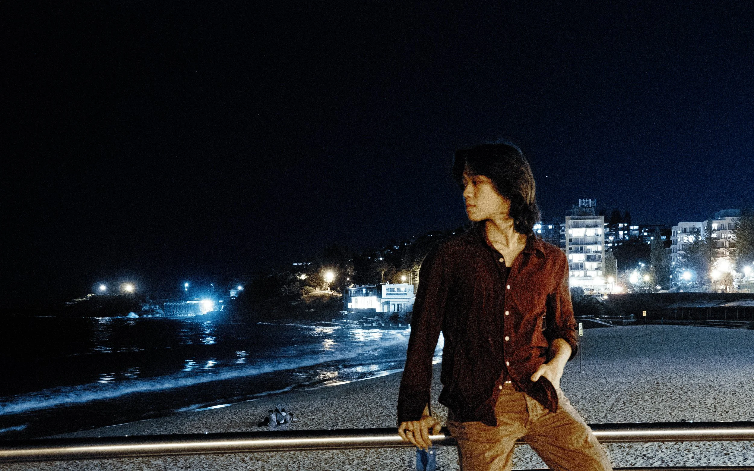 A young man with dark hair standing at the beach by a metal railing at night. He wears a red jacket and brown pants, looking to his right. The background shows a row of modern buildings illuminated against the night sky, with a few people sitting on the sand near the water. The sky is dark with some stars visible, and the water reflects city lights.