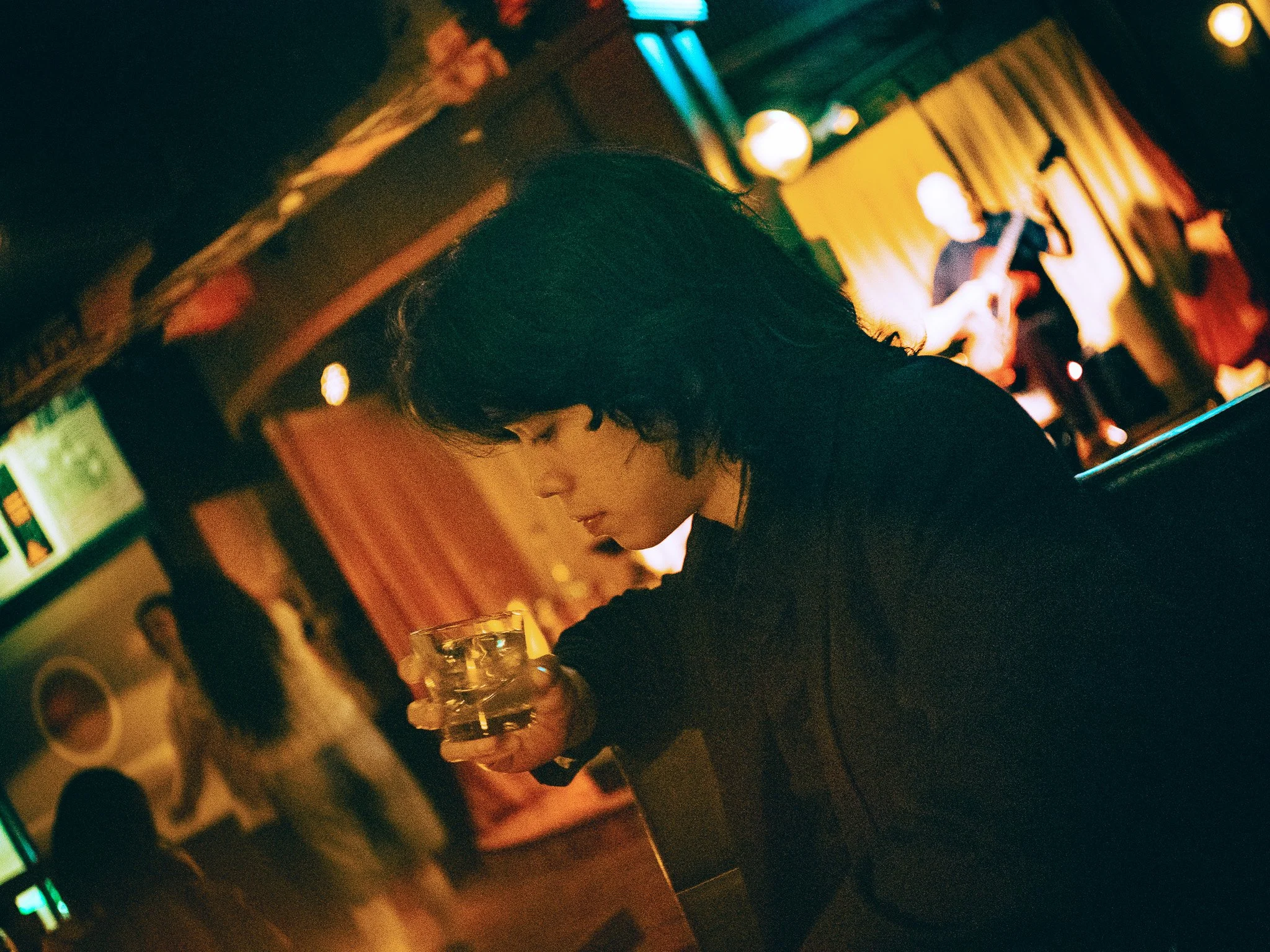 A person with short dark hair and a black shirt is sitting in a dimly lit bar with colorful lights and a musician playing guitar in the background.