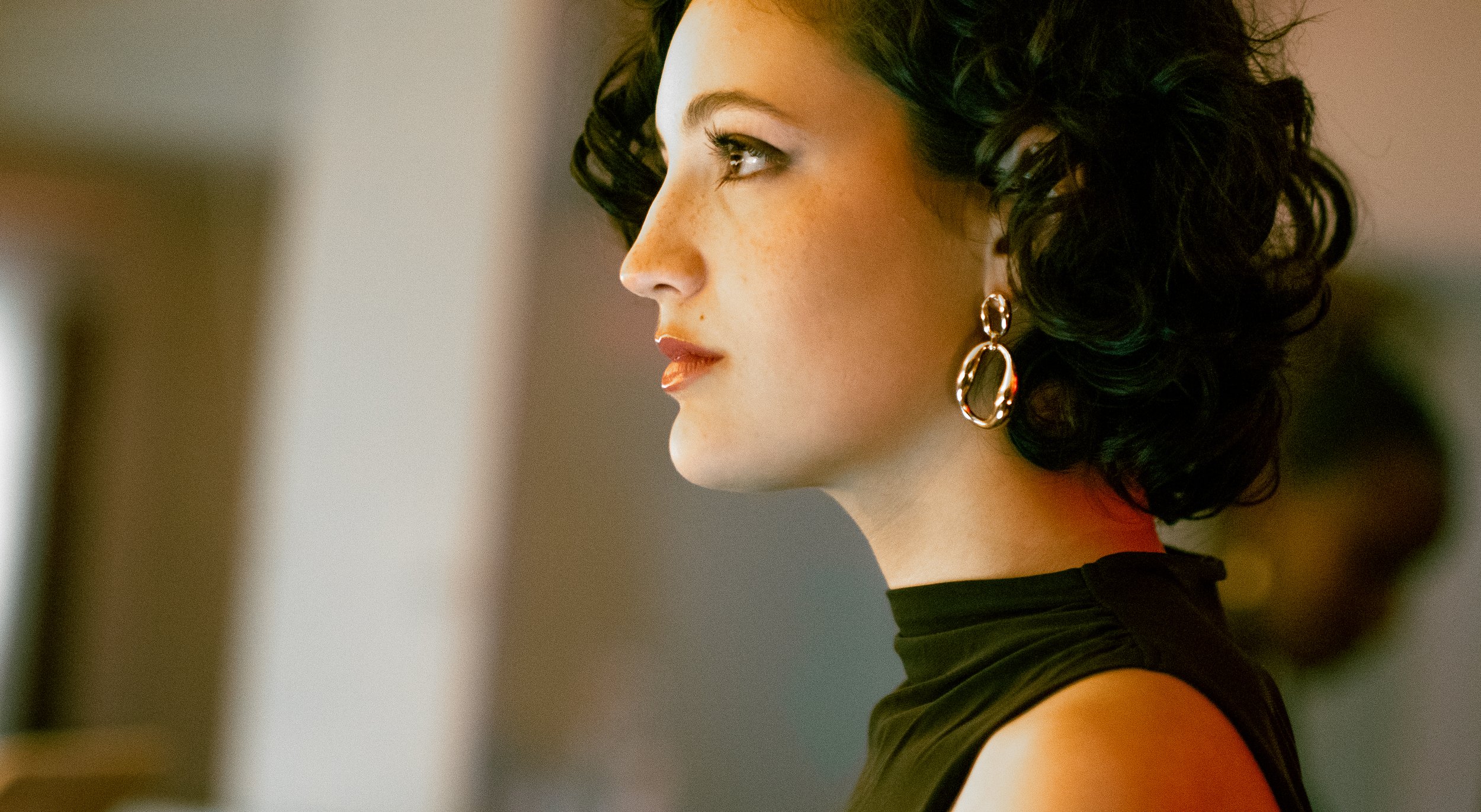 A young woman with curly dark hair, wearing drop earrings and a black turtleneck, looking to the side with a neutral expression.