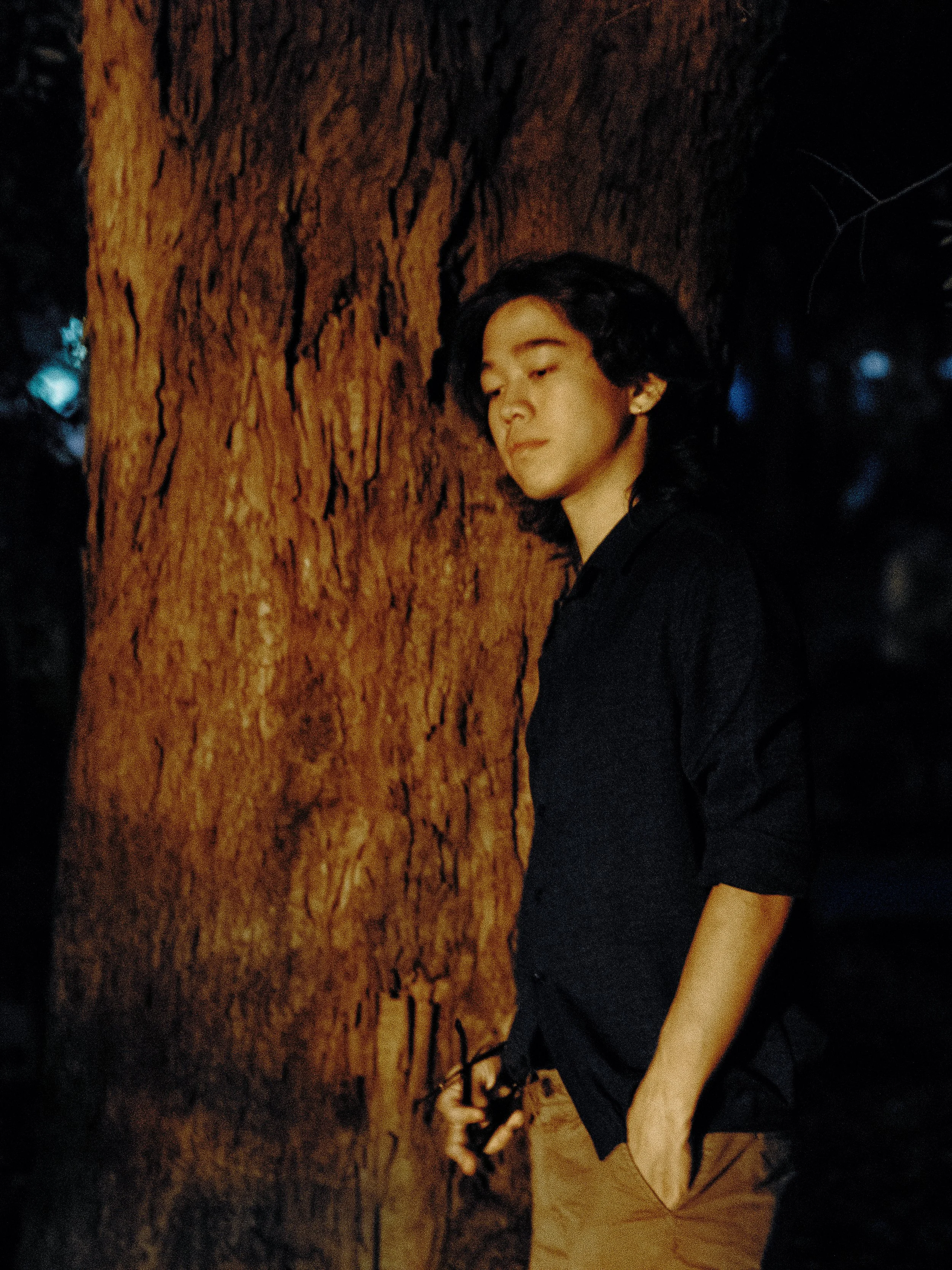 A young man with medium-length dark hair leaning against a large textured tree trunk at nighttime, wearing a black shirt and khaki pants, with his eyes closed and holding a black leash in his right hand.