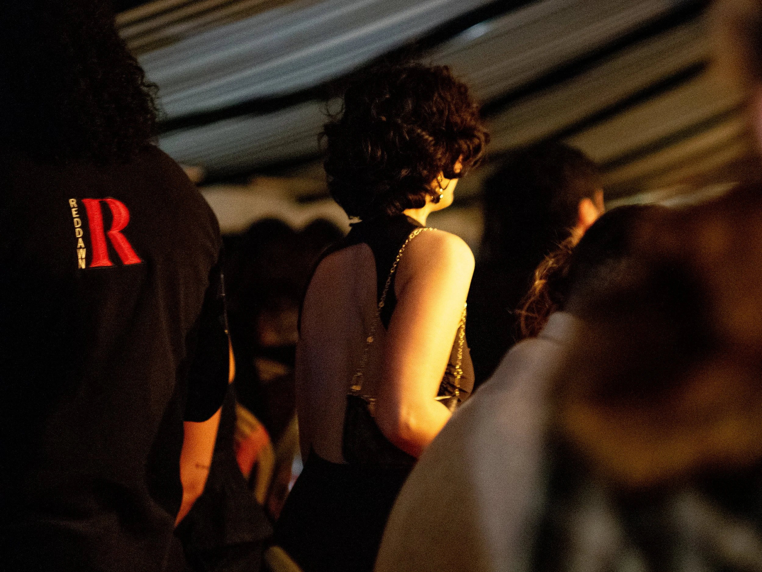 People standing in a line at an event, with one person wearing a black top with gold chains and a person wearing a black shirt with a red letter R on the sleeve.