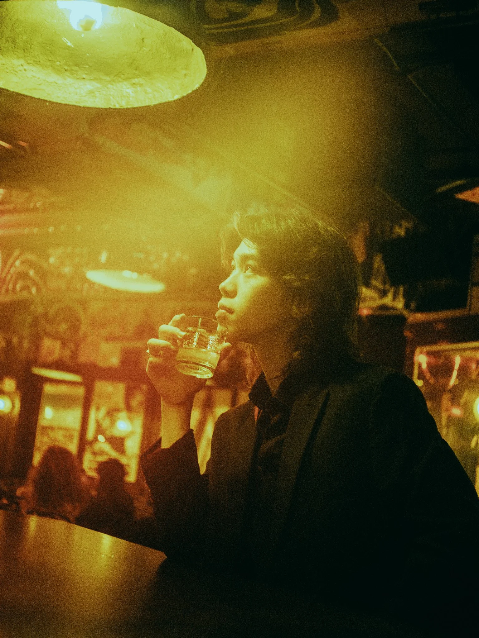 A young man with short dark hair is holding a glass of drink and looking to the left, in a dimly lit setting with warm yellow and orange lighting, possibly a bar or a restaurant.