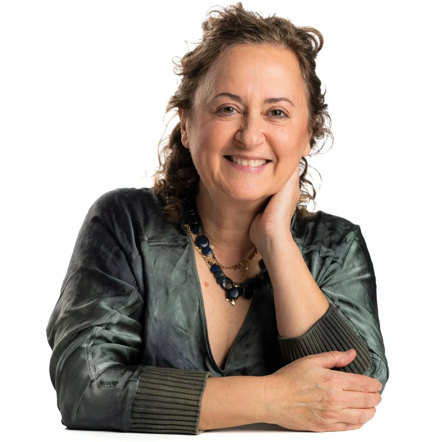 Mujer de cabello rizado, con ropa de tono gris, usa collar y pulsera, sonriente, con mano en la mejilla, en fondo blanco.