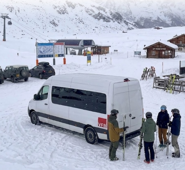 A group of four skiers in winter gear gathered around a white van in a snowy mountain ski resort. Ski equipment can be seen, along with a ski lift and small wooden buildings in the background.