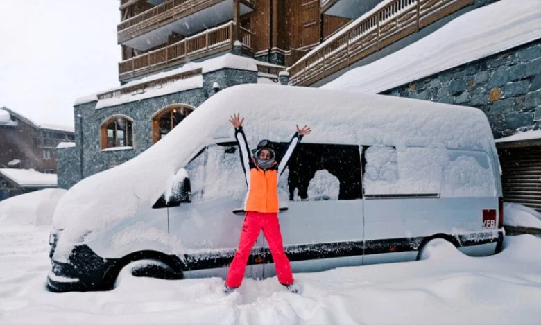 A person standing in front of a snow-covered van with arms raised, on a snowy day in a residential area.