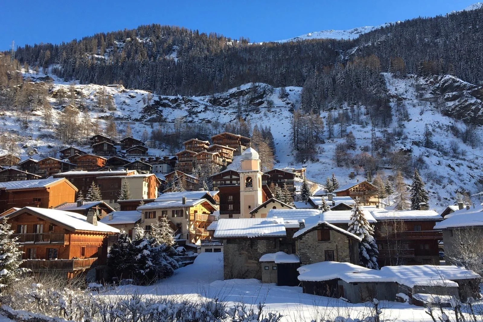 A snowy mountain village with wooden chalets and a church, surrounded by snow-covered trees and mountains under a clear blue sky.