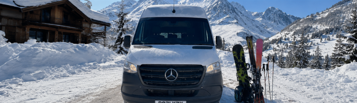 A white Mercedes-Benz van parked on a snowy mountain road, next to skis and ski poles, with snow-covered mountains and trees in the background.