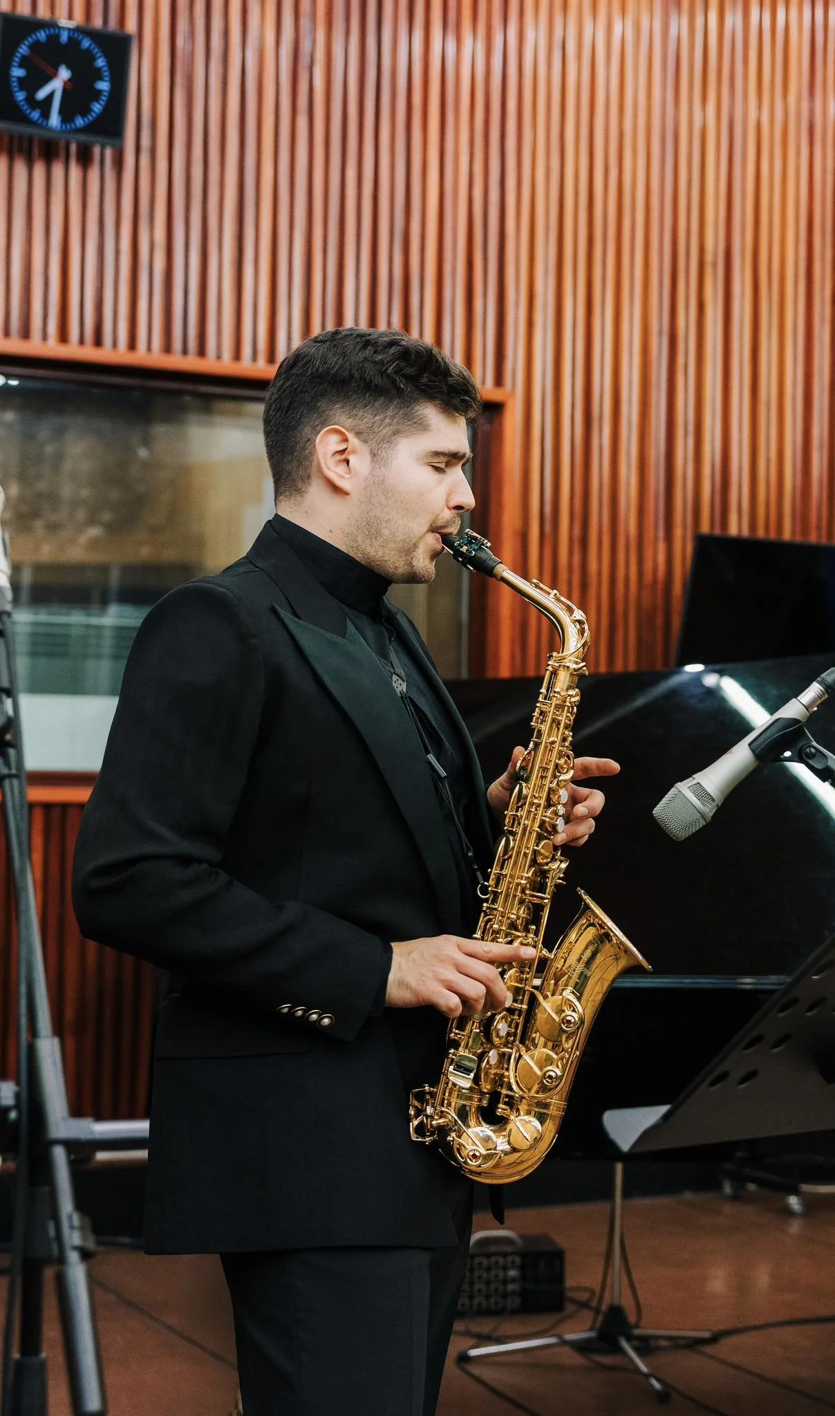 Ein Mann spielt Saxophon in einem Tonstudio, mit einem Klavier im Hintergrund und einer Mikrofonaufnahme. Die Wände sind mit Holz verkleidet, und eine Uhr ist sichtbar.