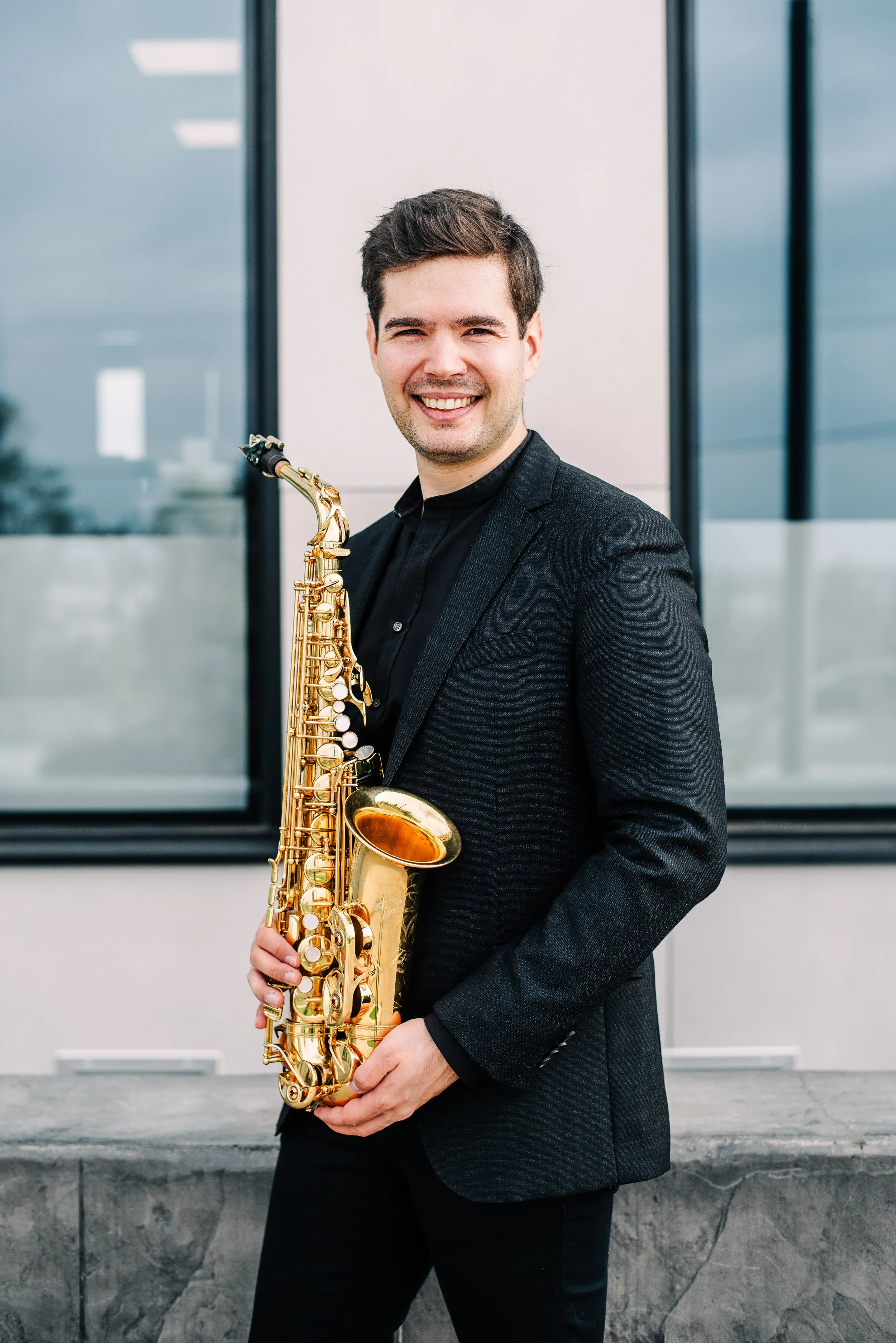 Ein junger Mann trägt einen schwarzen Anzug und hält ein goldfarbenes Saxophon vor einem modernen Gebäude mit großen Glasfenstern.