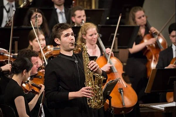 Ein Orchester spielt, ein Musiker im Vordergrund spielt Saxophon, andere Musiker spielen Violinen im Hintergrund.