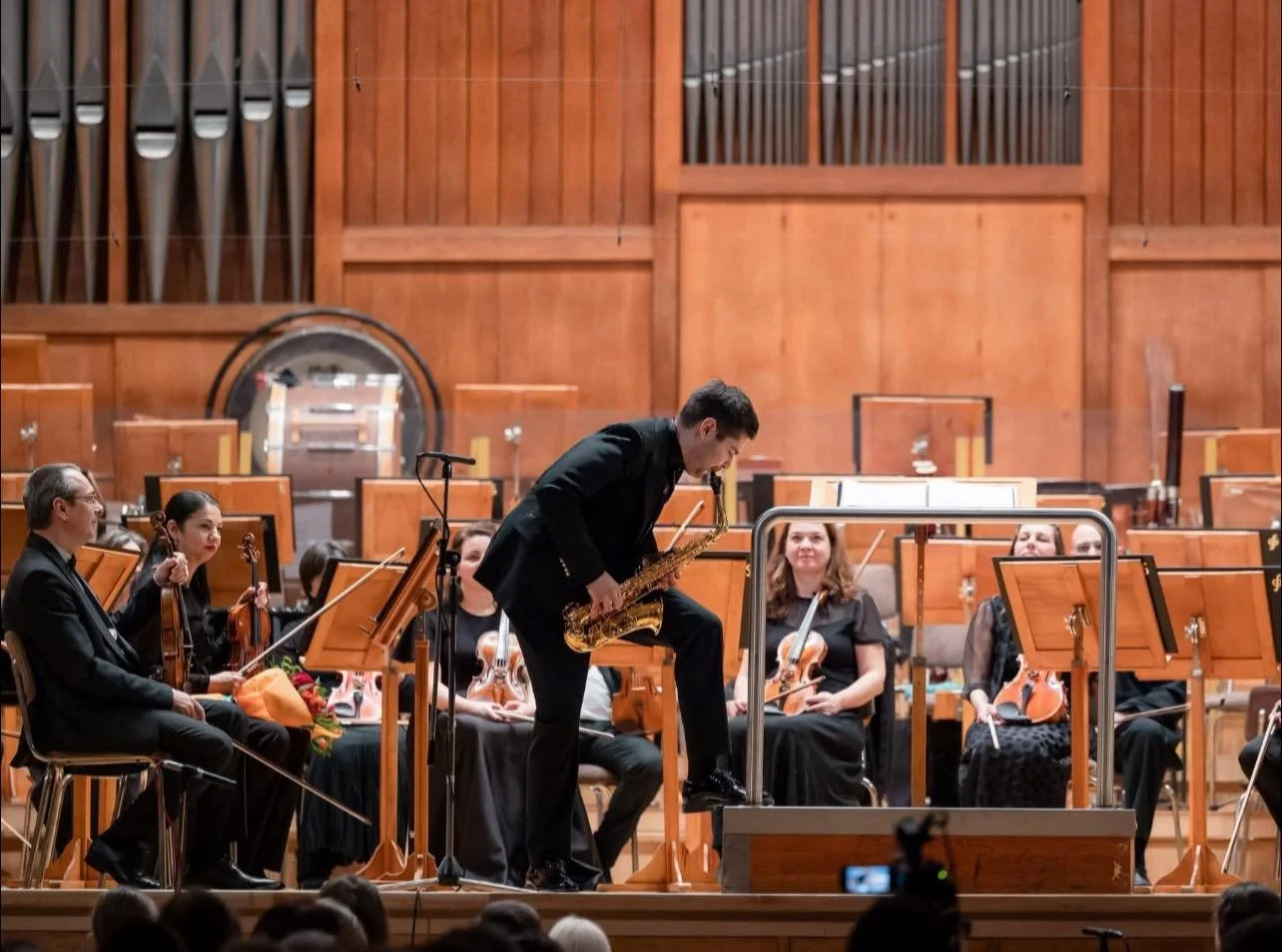 Ein Orchester auf der Bühne, ein Musiker spielt Saxophon, andere Musiker mit Violinen sitzen im Hintergrund.