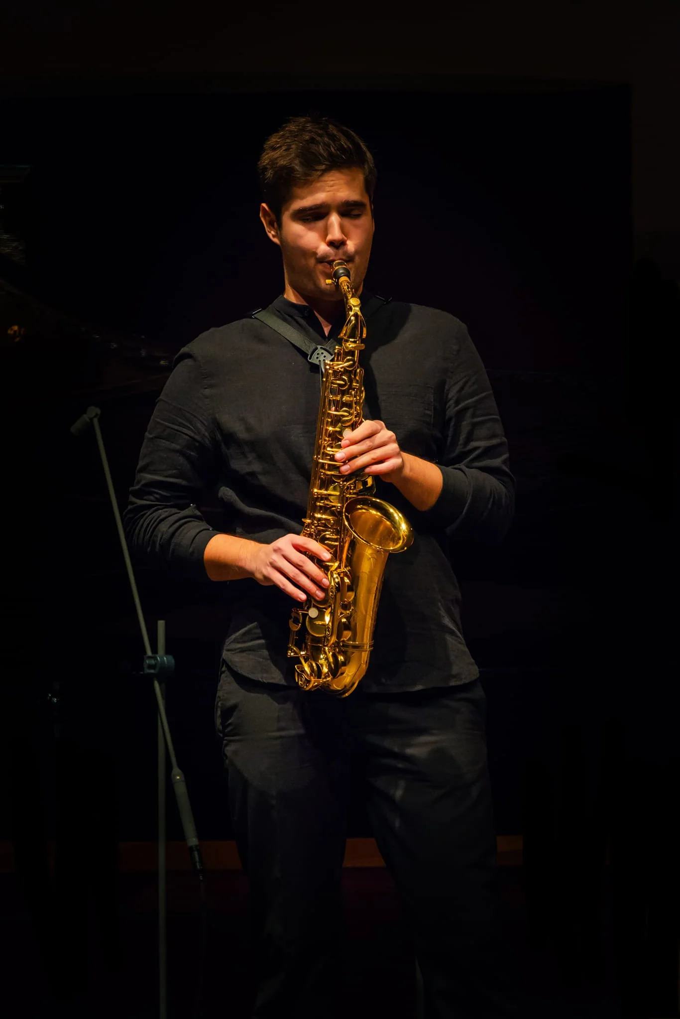 Junger Mann spielt Saxophon auf einer Bühne, dunkler Hintergrund, konzentriert beim Musizieren.