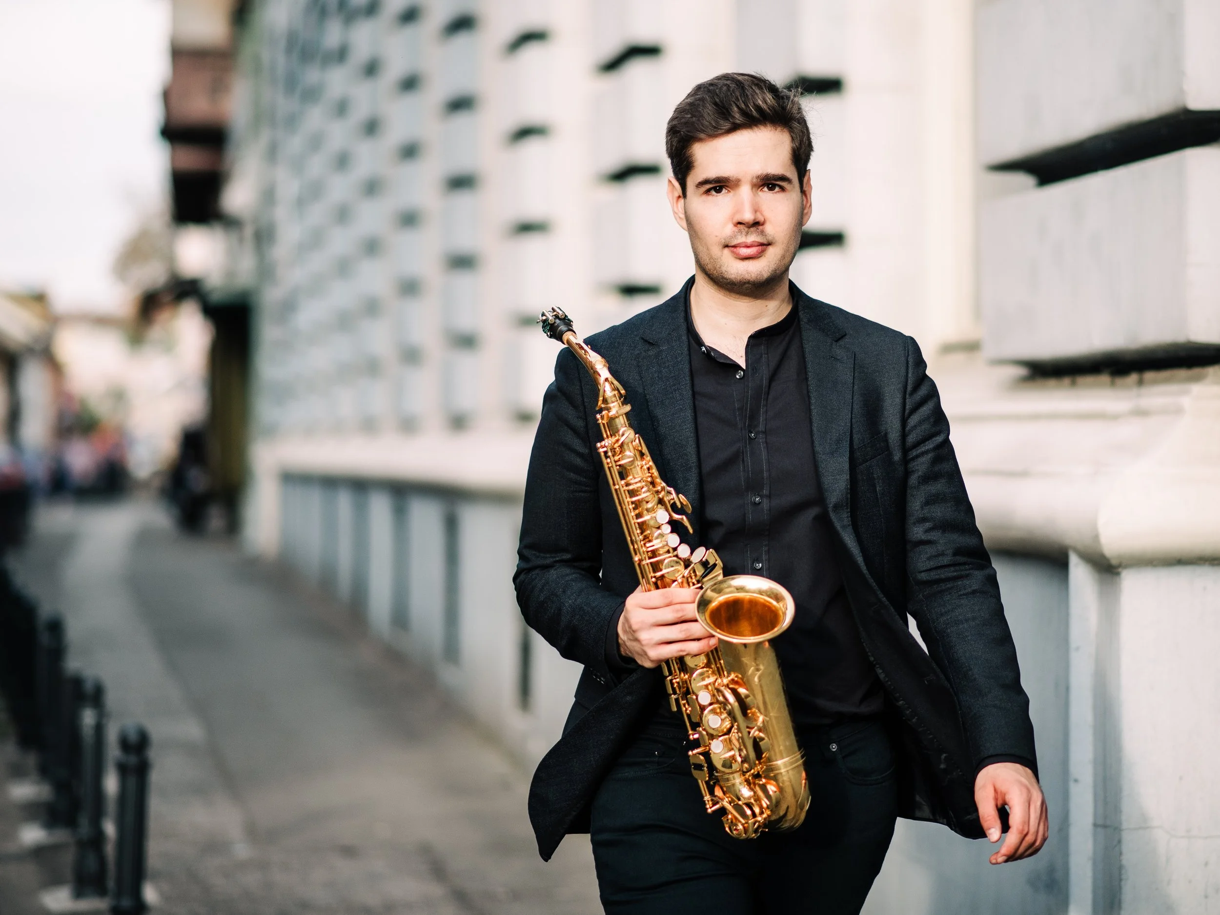 Ein junger Mann in dunkler Kleidung, der auf einer Straße in einer Stadt mit modernen Gebäuden einen goldenen Saxophon hält.