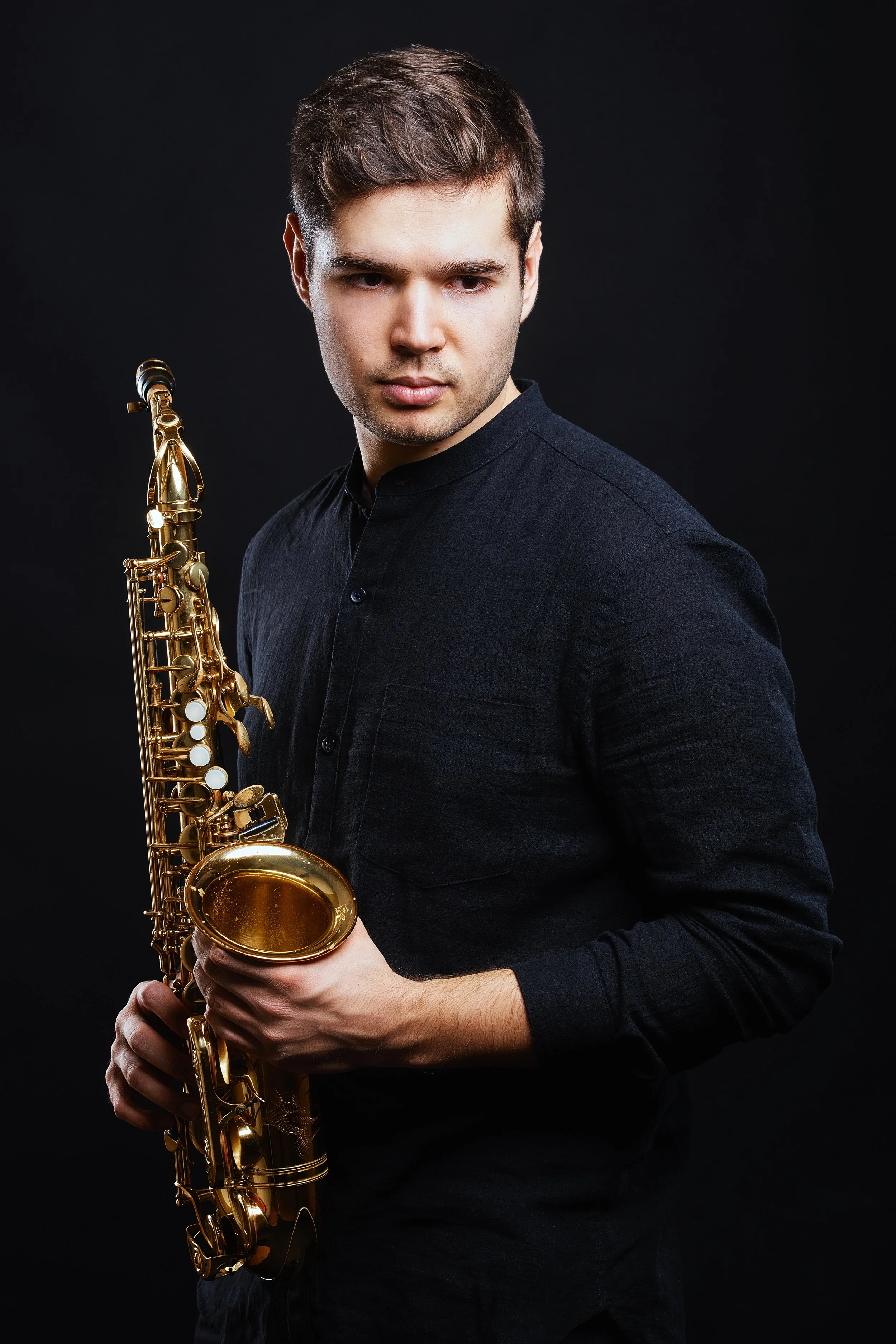 Junger Mann mit dunklem Hemd hält Saxophon vor einem schwarzen Hintergrund.